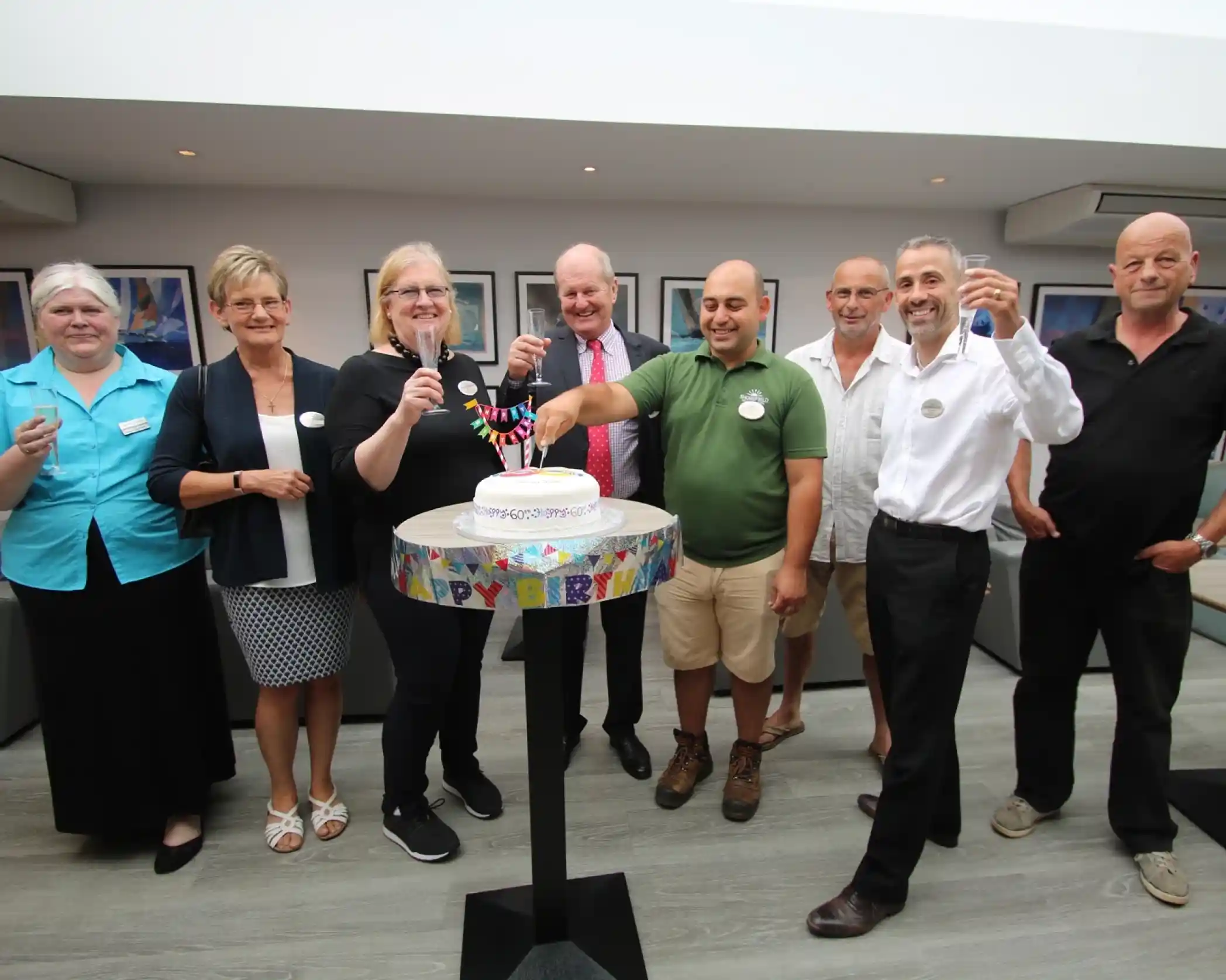 This image shows a group of eight people celebrating a 60th birthday. They are standing around a cake on a tall stand that has a colorful 