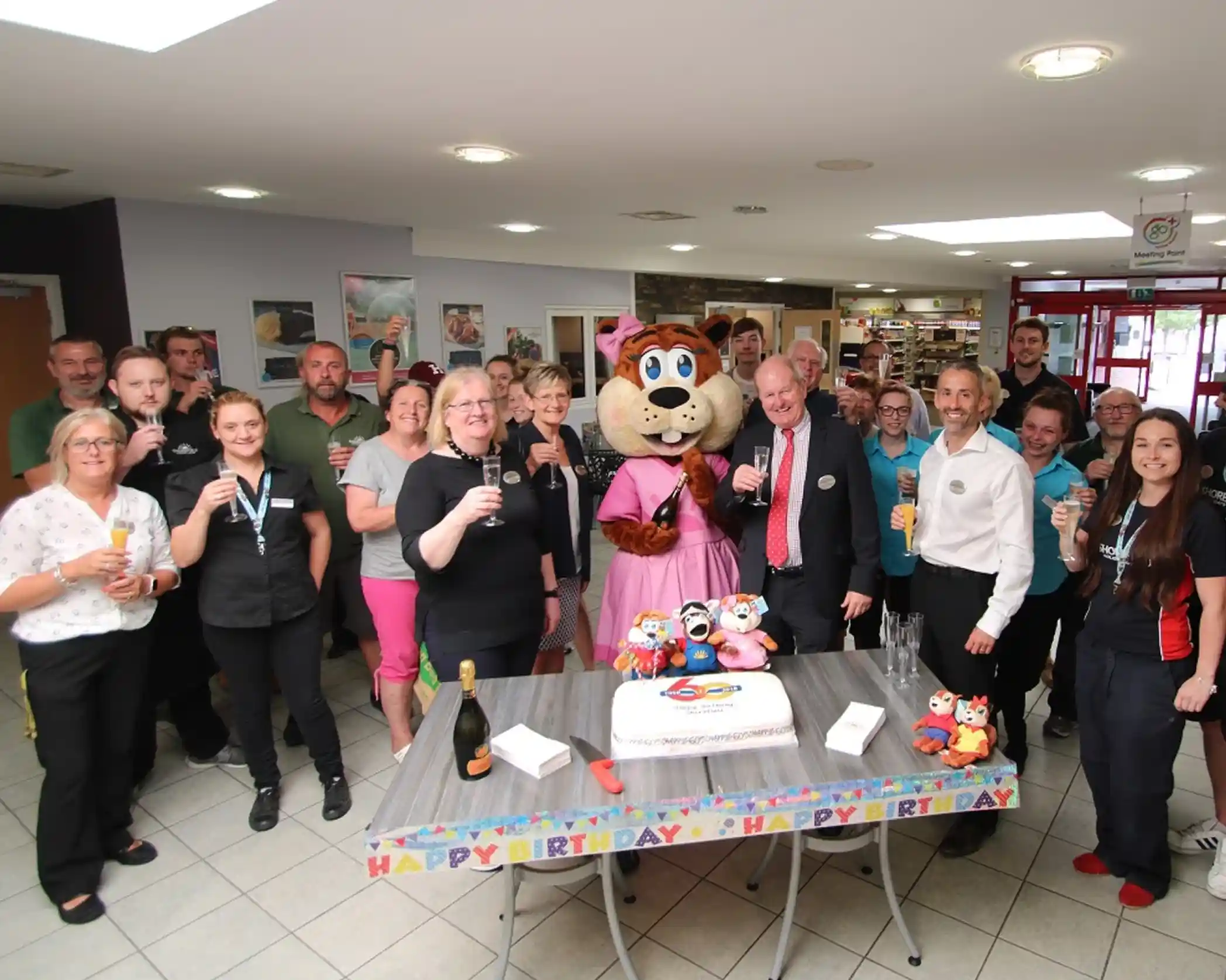 This image depicts a group of people celebrating a 60th birthday event. They are gathered around a table with a cake that has 