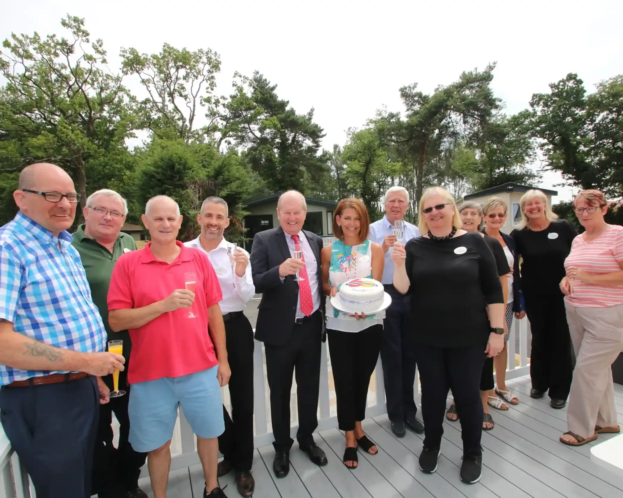 This image shows a group of twelve people standing outdoors on a deck, celebrating an event. Most of them are holding drinks, and one woman near the center is holding a cake decorated with a 