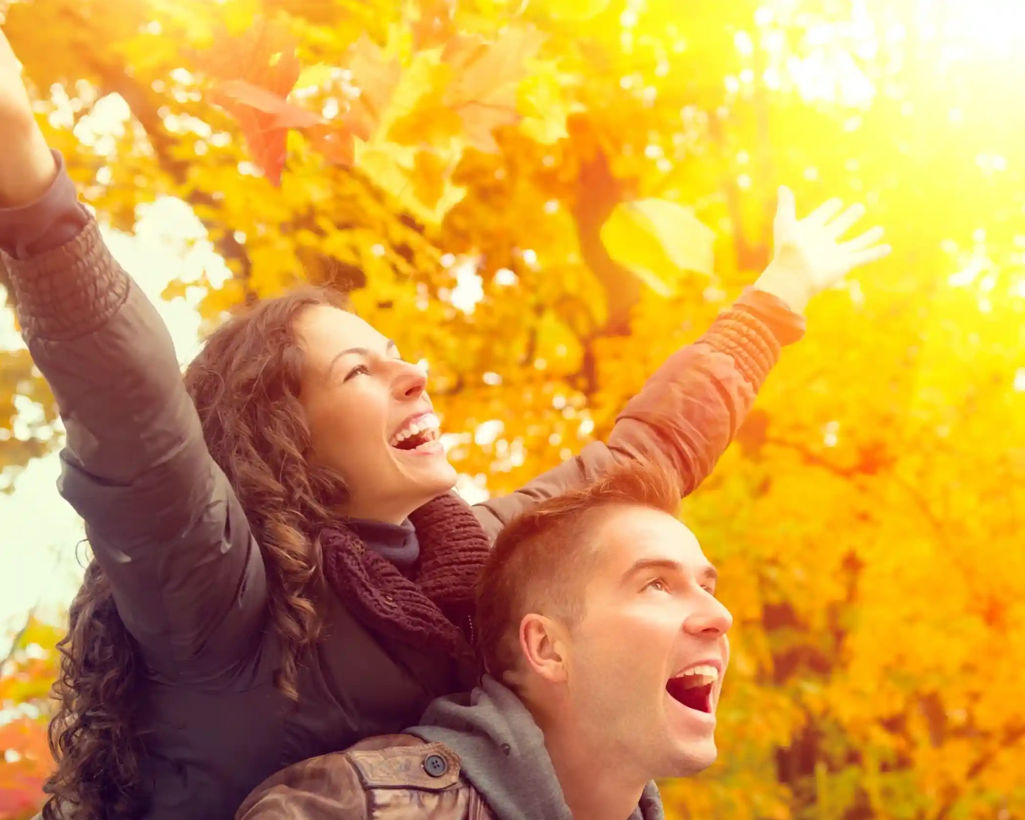 A couple laughing and smiling in the forest. The lady has both hands up in joy and wonder at the autumn leaves around them whilst piggybacking on her partner's back. Lighting is soft and autumnal.