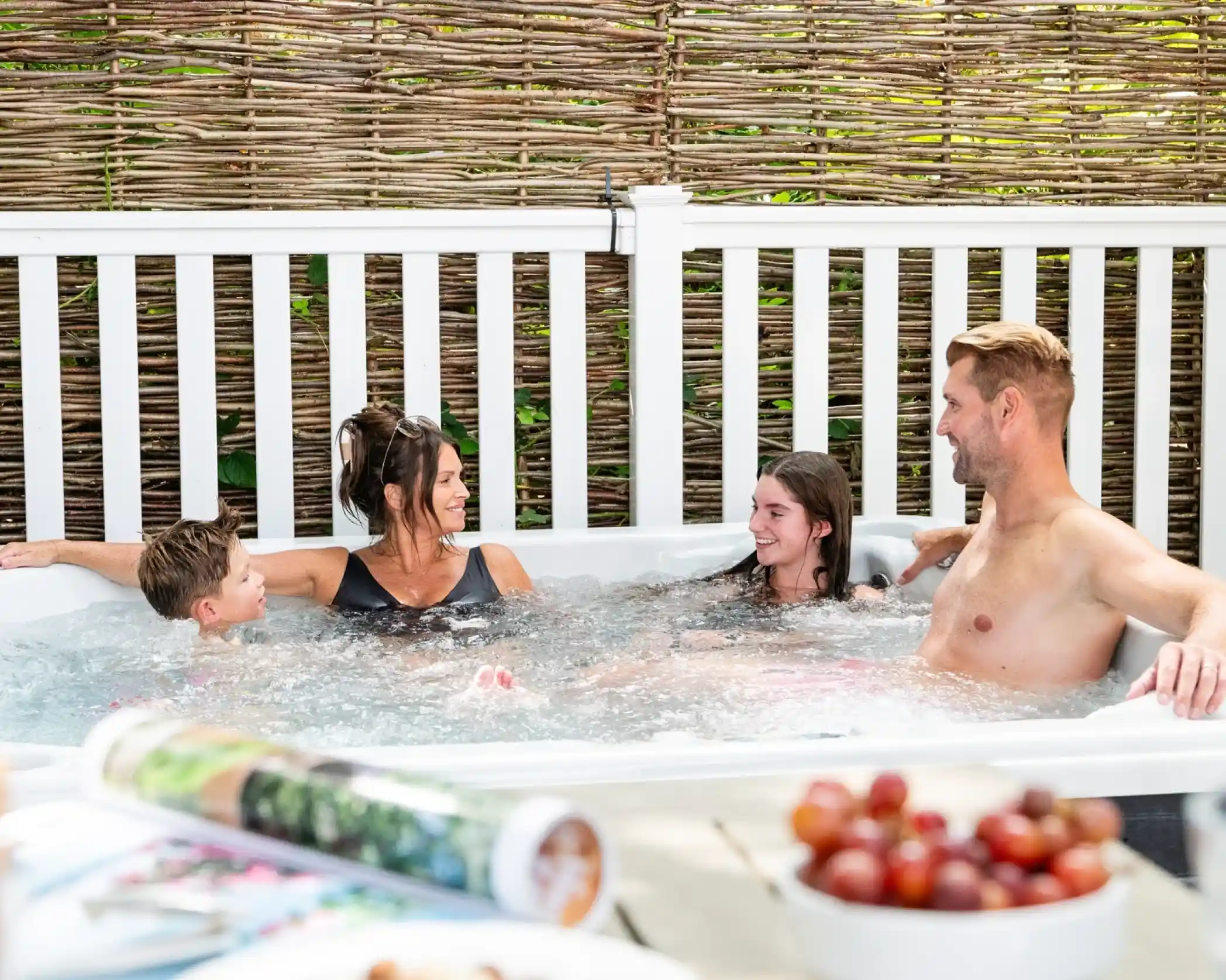 A family of four, husband and wife and a girl and boy, are sitting in a hot tub with a private weaved fence behind them and some food and a magazine on a table out of focus in the foreground.