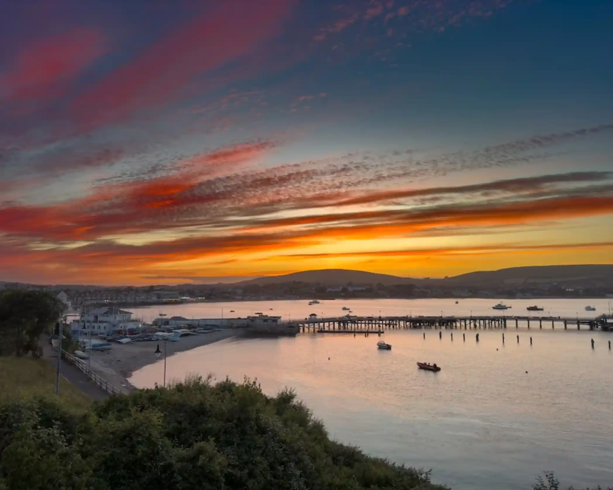 This image shows a stunning sunset over a peaceful coastal town. The vibrant sky is painted in hues of orange, pink, and purple, reflecting off the calm waters. A pier stretches into the sea, with several boats docked along it, creating a tranquil, scenic view. The surrounding landscape, with hills in the distance and buildings near the shore, further enhances the beauty of the scene. The golden light of the sunset casts a serene glow on the entire bay, offering a perfect moment of calm by the sea.
