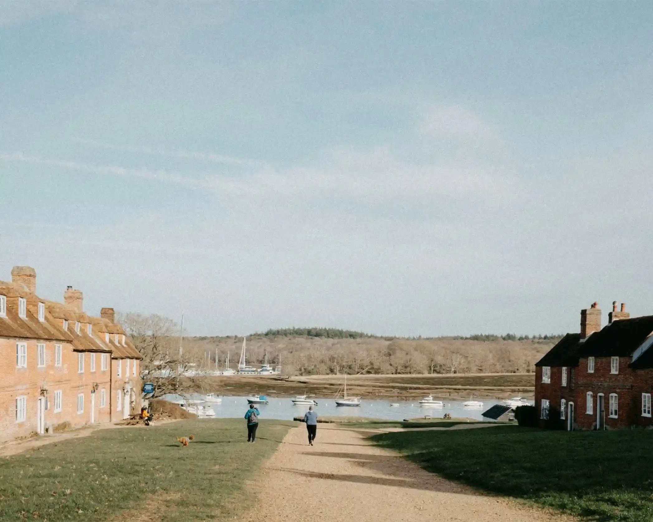 A gravel path leads between red-brick houses towards a calm waterway, where boats are anchored. Two people walk along the path, surrounded by trees and hills in the distance under a partly cloudy sky.