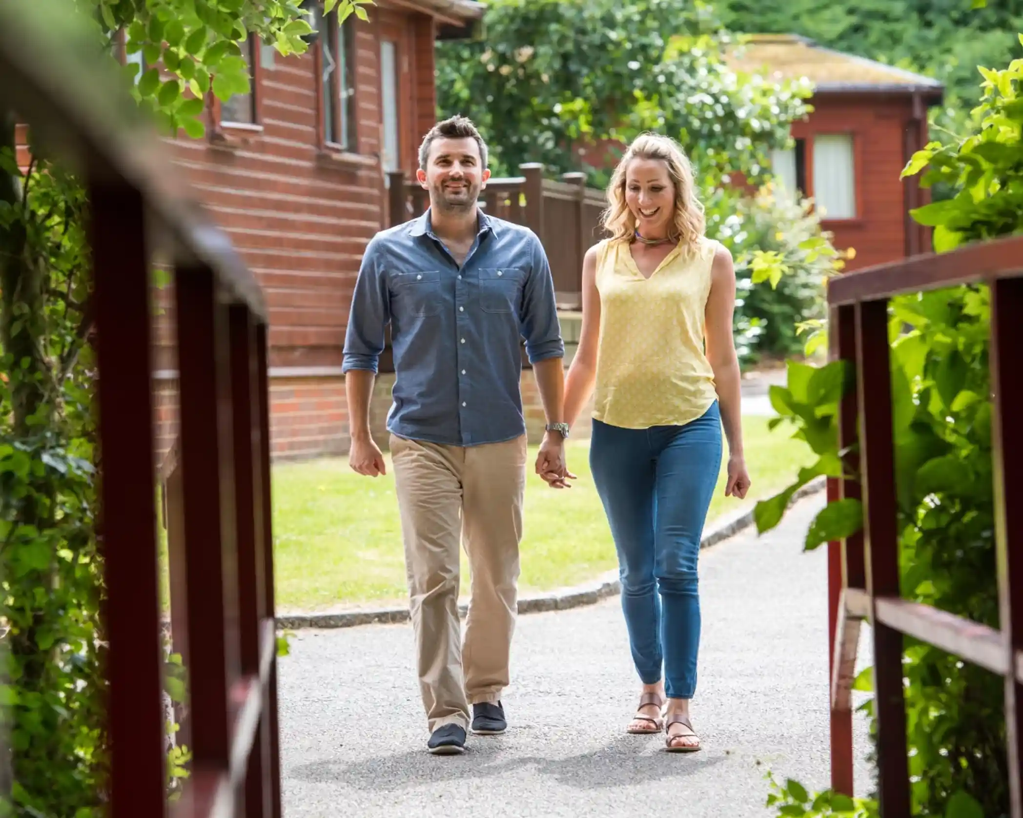Smiling couple walking hand-in-hand along a tree-lined path in a holiday park with wooden lodges and greenery on either side.