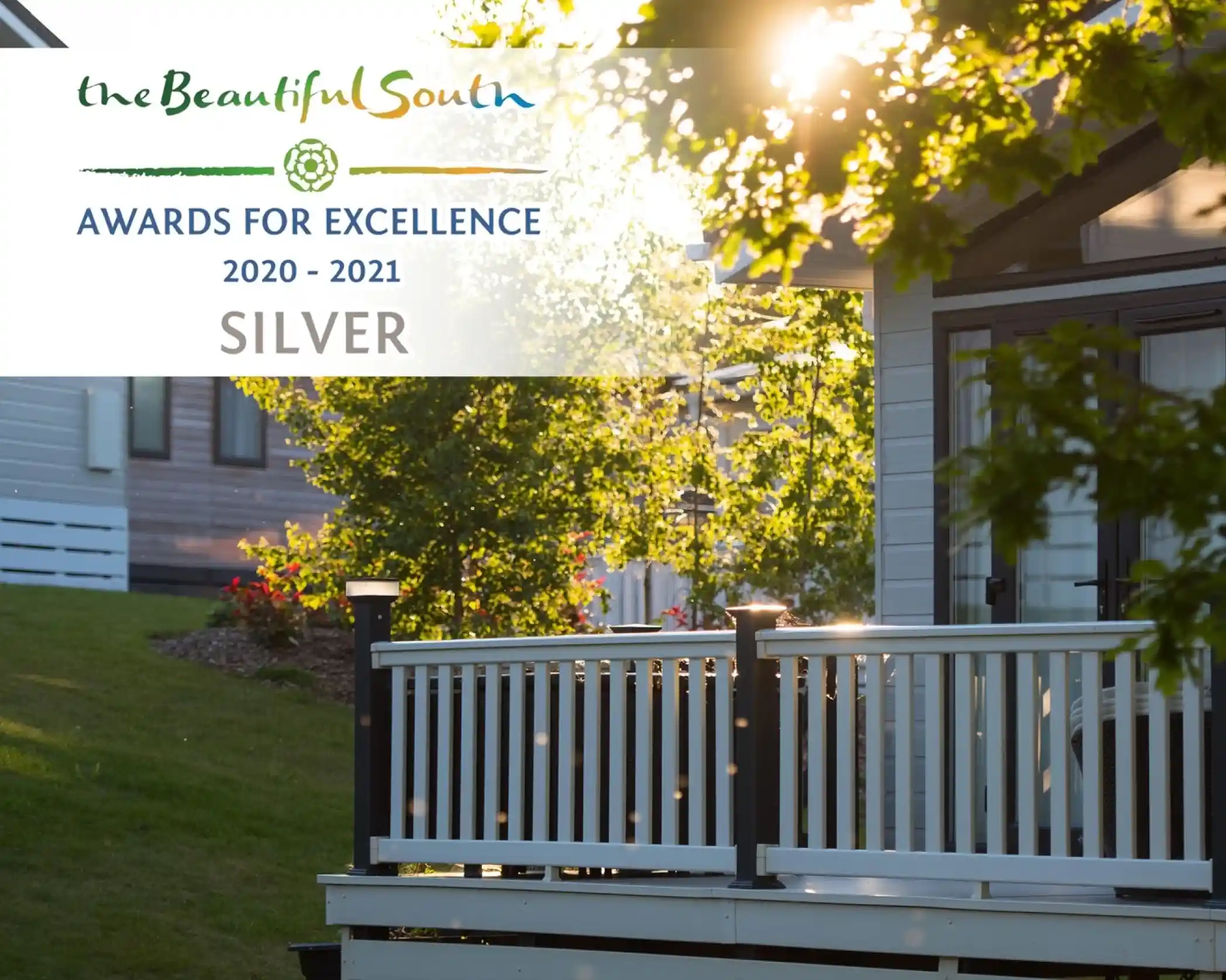 Holiday lodge park at sunset with modern static caravans, well-maintained landscaping, and a white wooden deck in the foreground. Overlaid is a 'Beautiful South Awards for Excellence 2020-2021 Silver' badge.