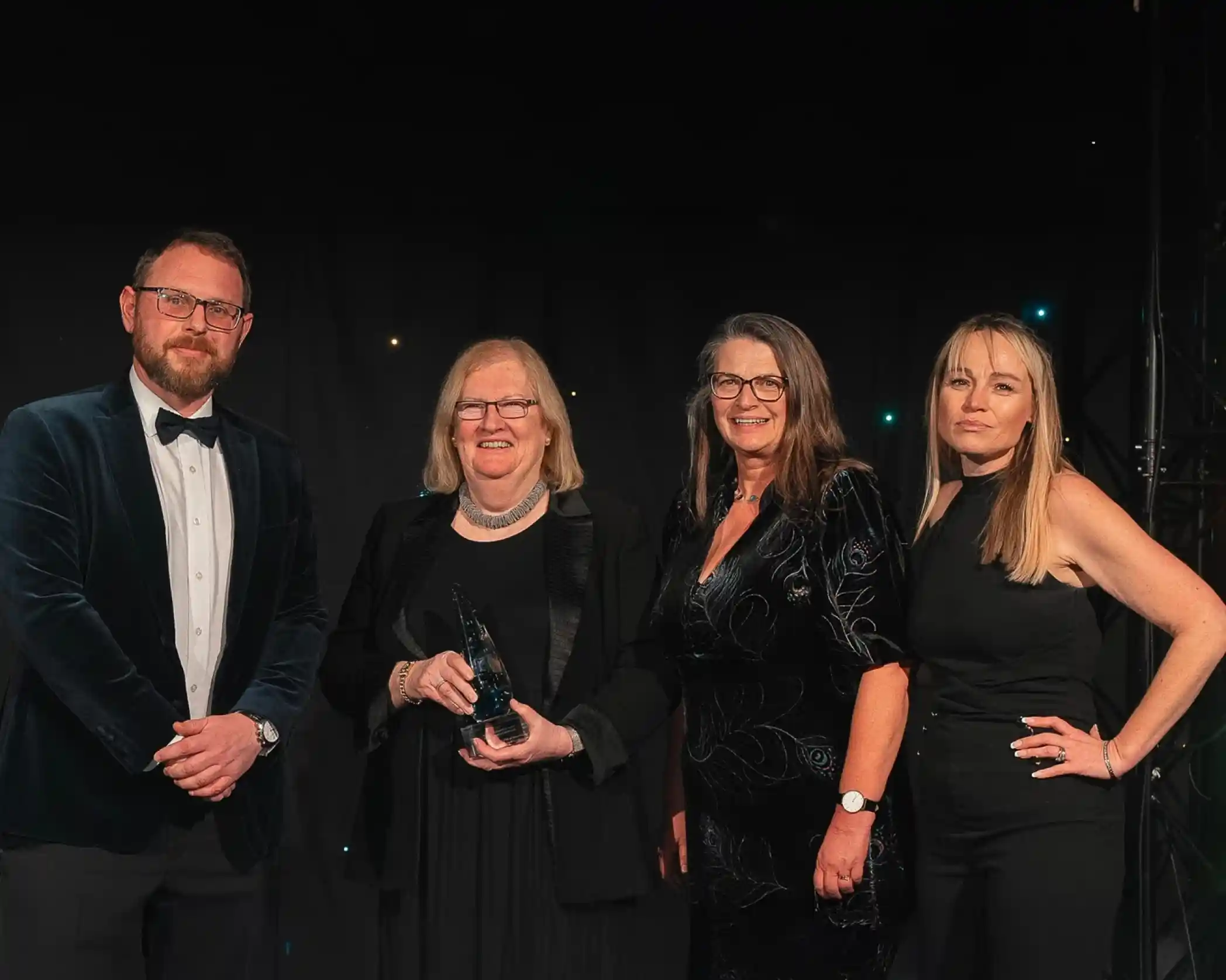 Four people dressed formally, standing together at an awards event. The second person from the left, an older woman, is holding a glass award and smiling. The backdrop is dark with small glowing lights, creating a celebratory atmosphere.