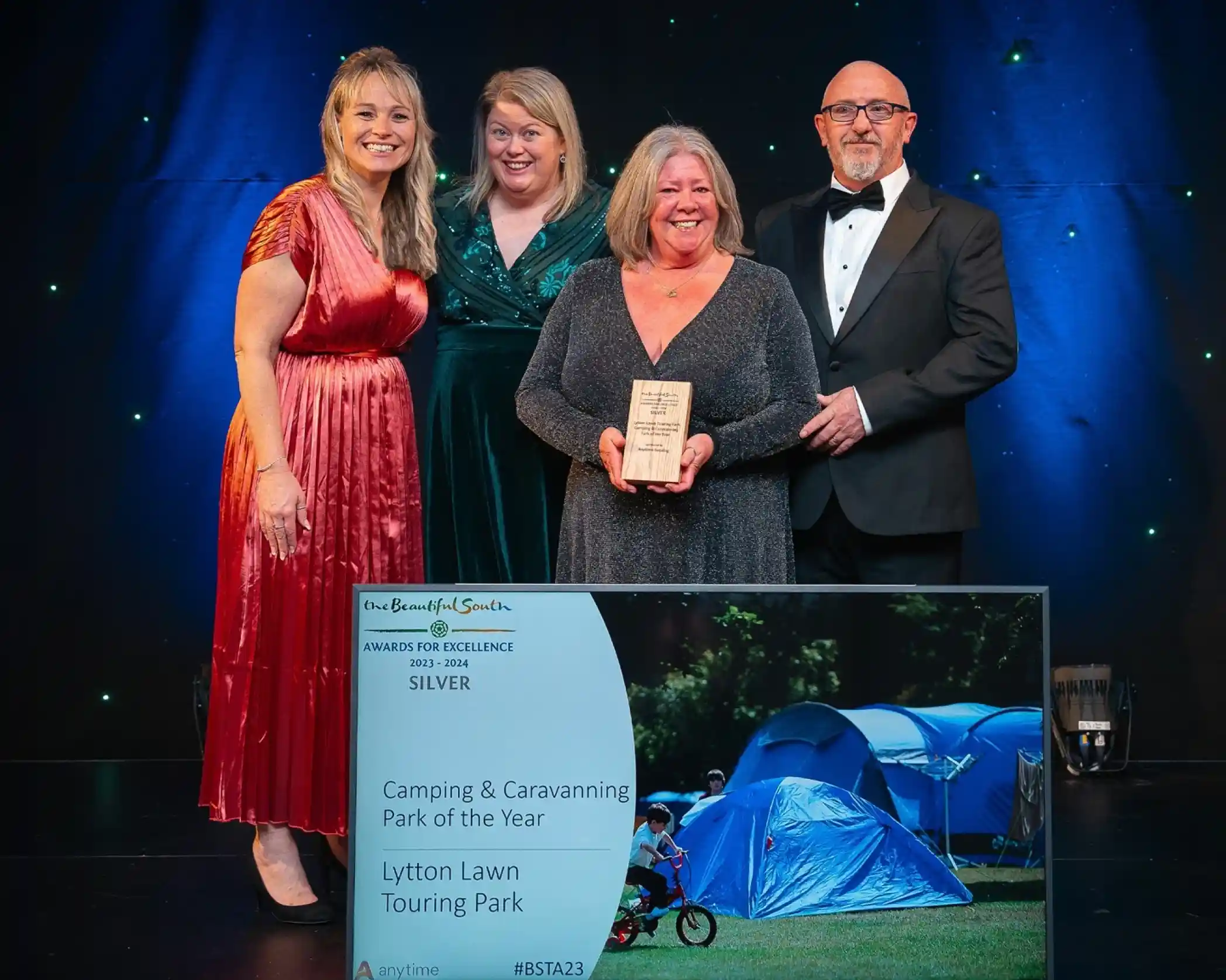 Four people, smiling and in formal attire, are stood against a dark backdrop with small lights, and the second person from the left, an older lady, is holding a wooden award. In front of them on the floor is a large framed award with an image of tents in a field with the words 'Camping & Caravanning Park of the Year - Lytton Lawn Touring Park'.