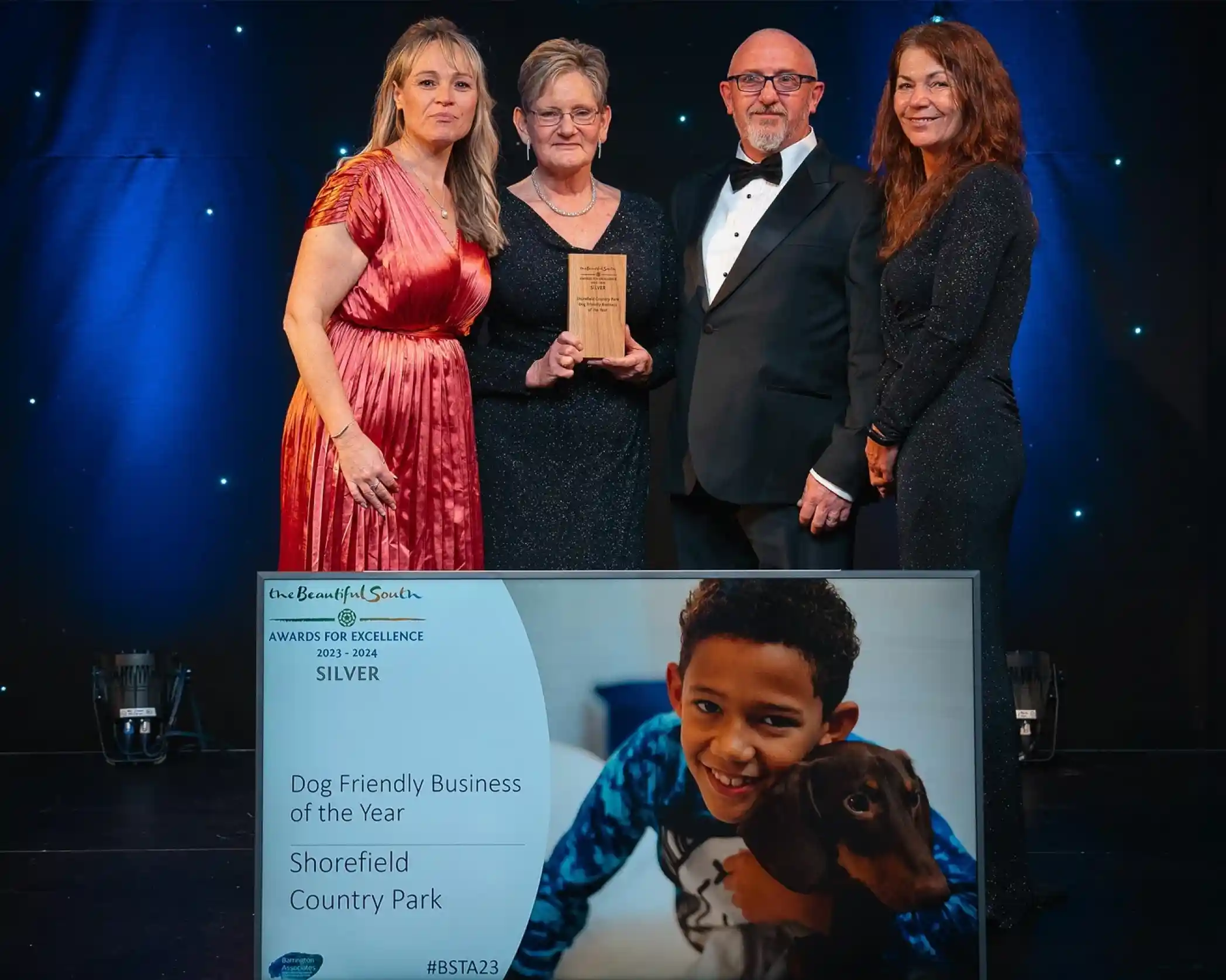 Four people, smiling and in formal attire, are stood against a dark backdrop with small lights, and the second person from the left, an older lady, is holding a wooden award. In front of them on the floor is a large framed award with an image of a young boy cuddling a dog with the words 'Dog Friendly Business of the Year - Shorefield Country Park'.