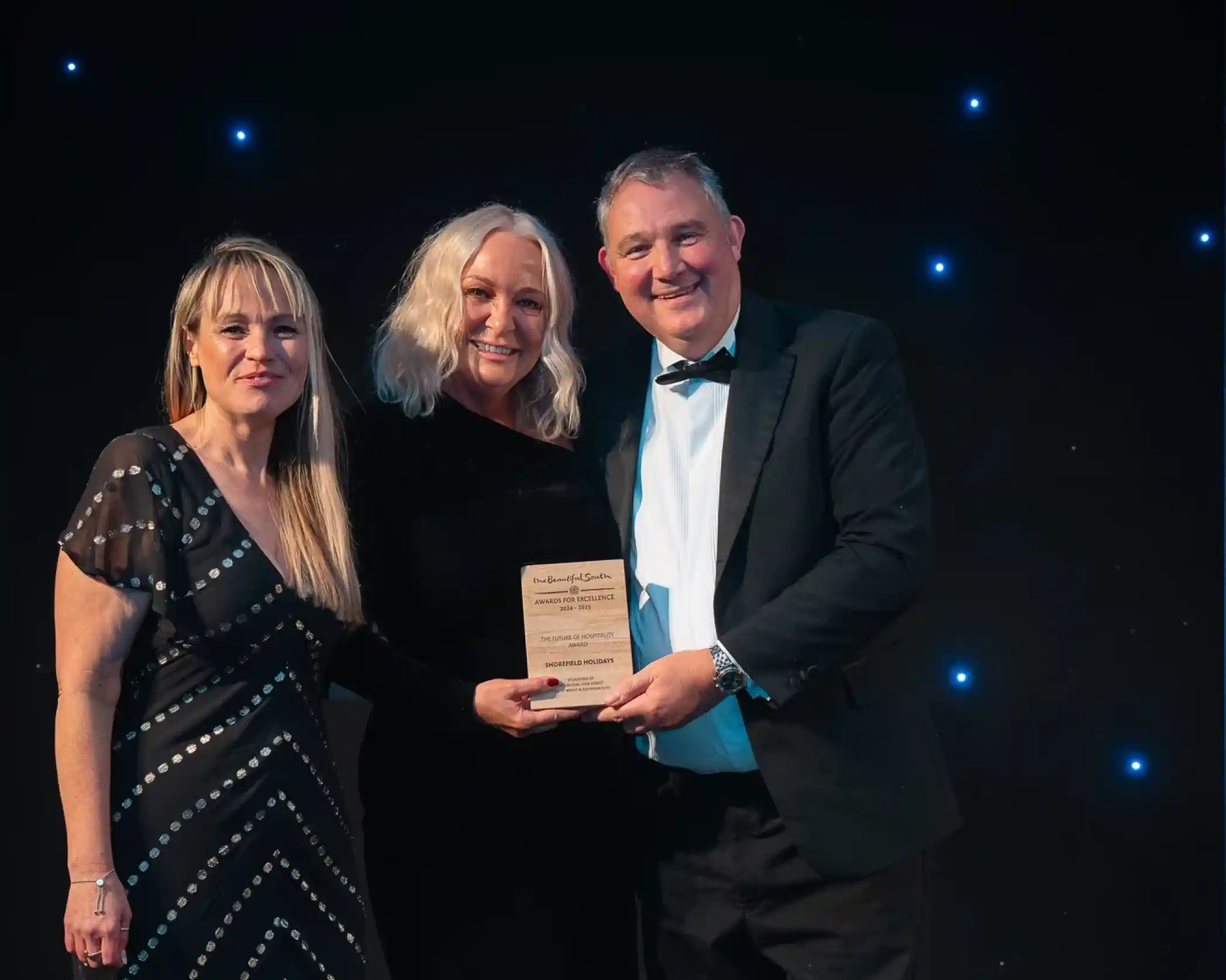 Three people dressed in formal attire, standing together at an awards event. The middle lady and the man on the right are both holding onto a wooden award together and smiling. The backdrop is dark with small glowing lights, creating a celebratory atmosphere.