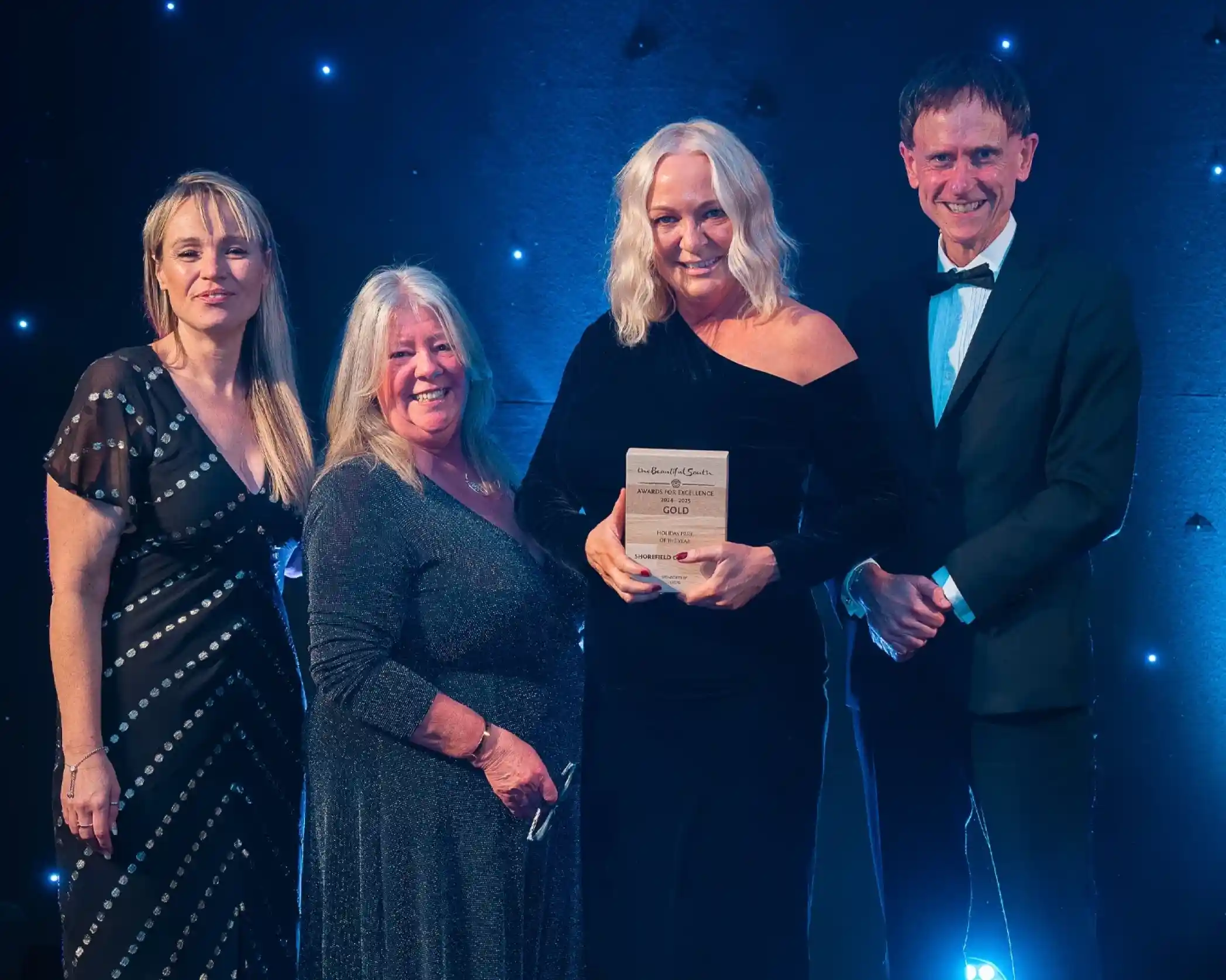 Four people dressed formally, standing together at an awards event. The second person from the right, a woman, is holding a wooden award and smiling. The backdrop is dark blue with small glowing lights, creating a celebratory atmosphere.