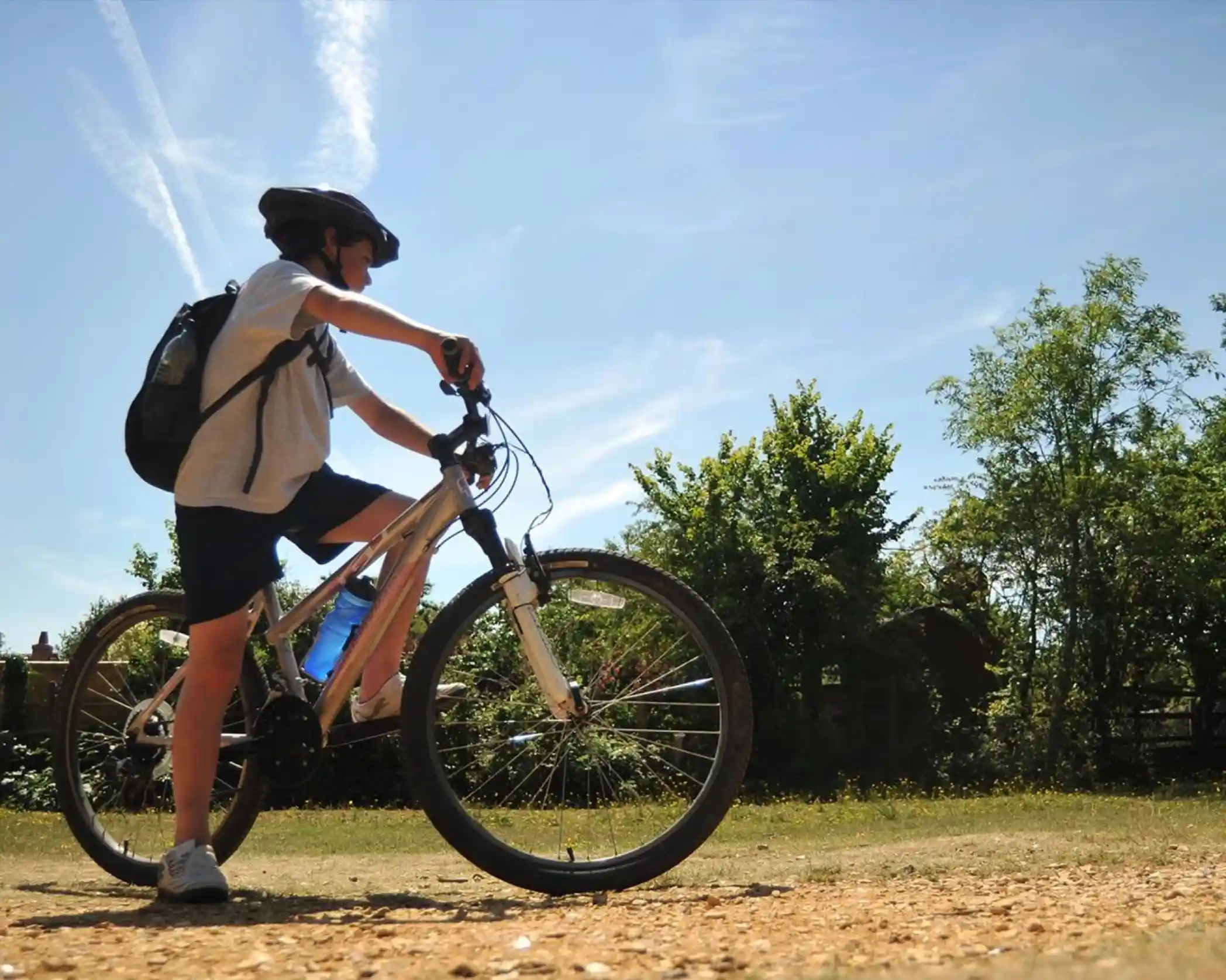 A young boy is on a bike, stood with one foot on the ground and one foot ready to pedal, wearing a rucksack and helmet. He is in the forest on a gravel path with trees in the distance.