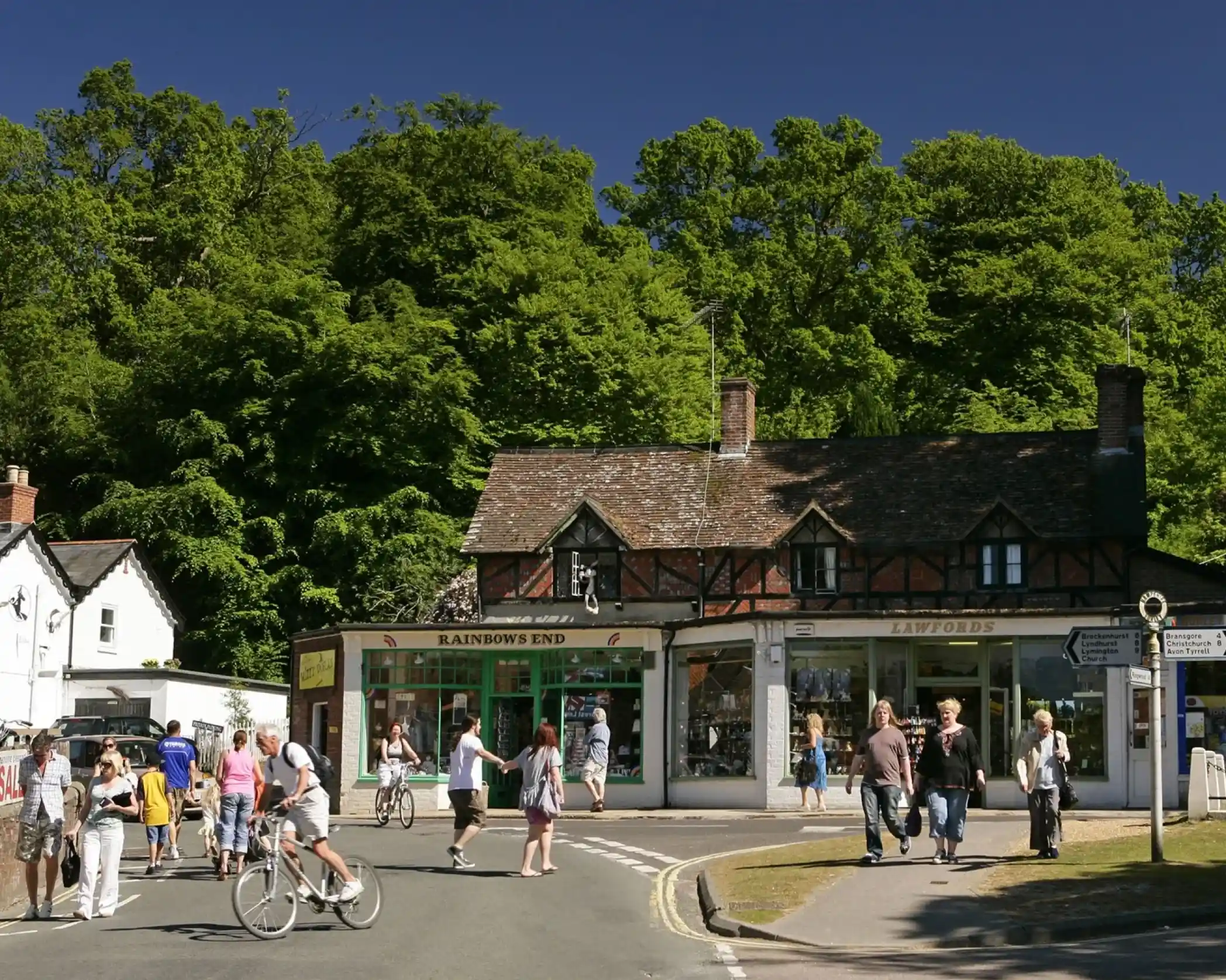 People walking and cycling through Burley village in the New Forest, with shops and trees in the background.