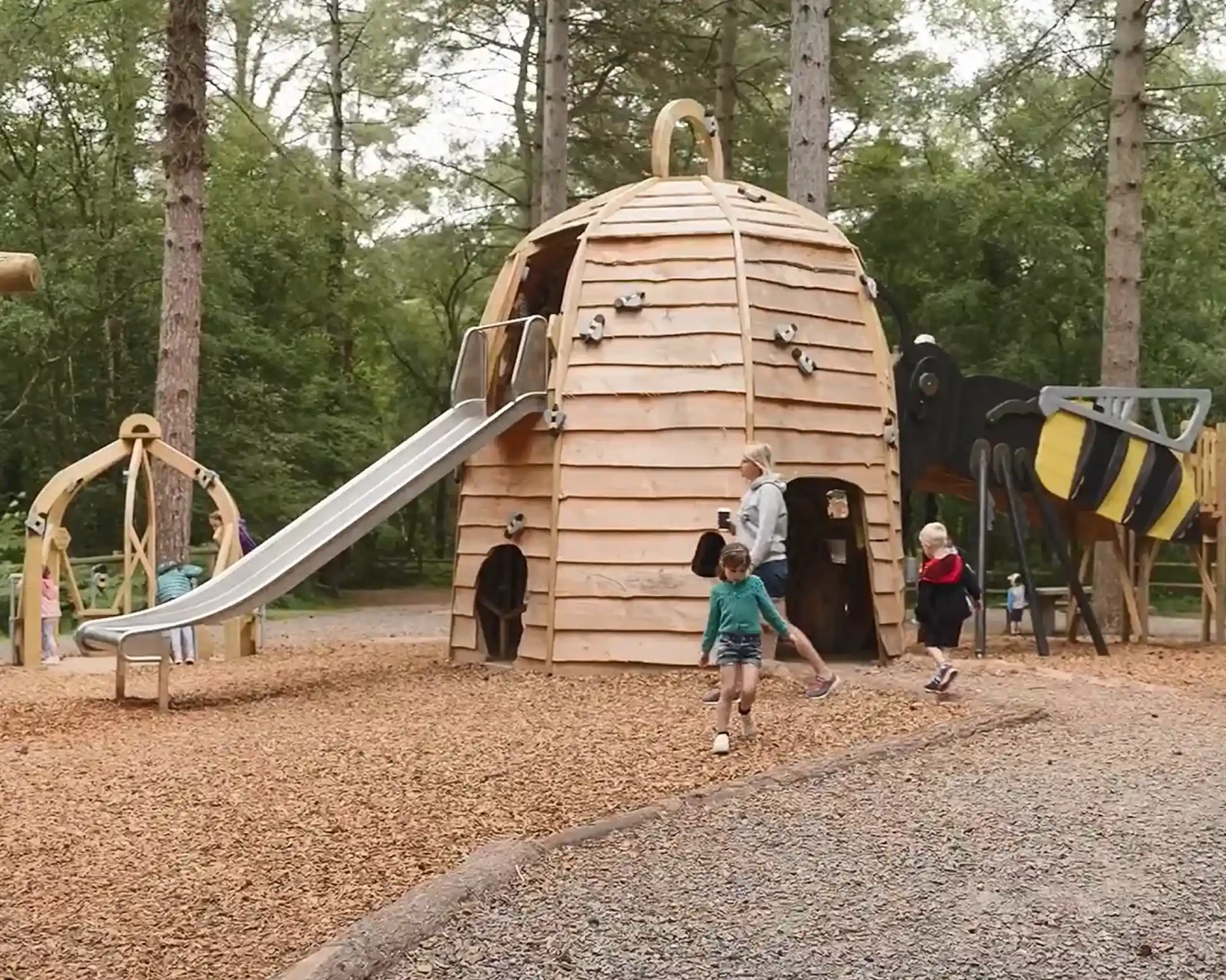 A wooden play structure resembling a beehive features a large slide, surrounded by children playing in a forested area with soft ground cover.