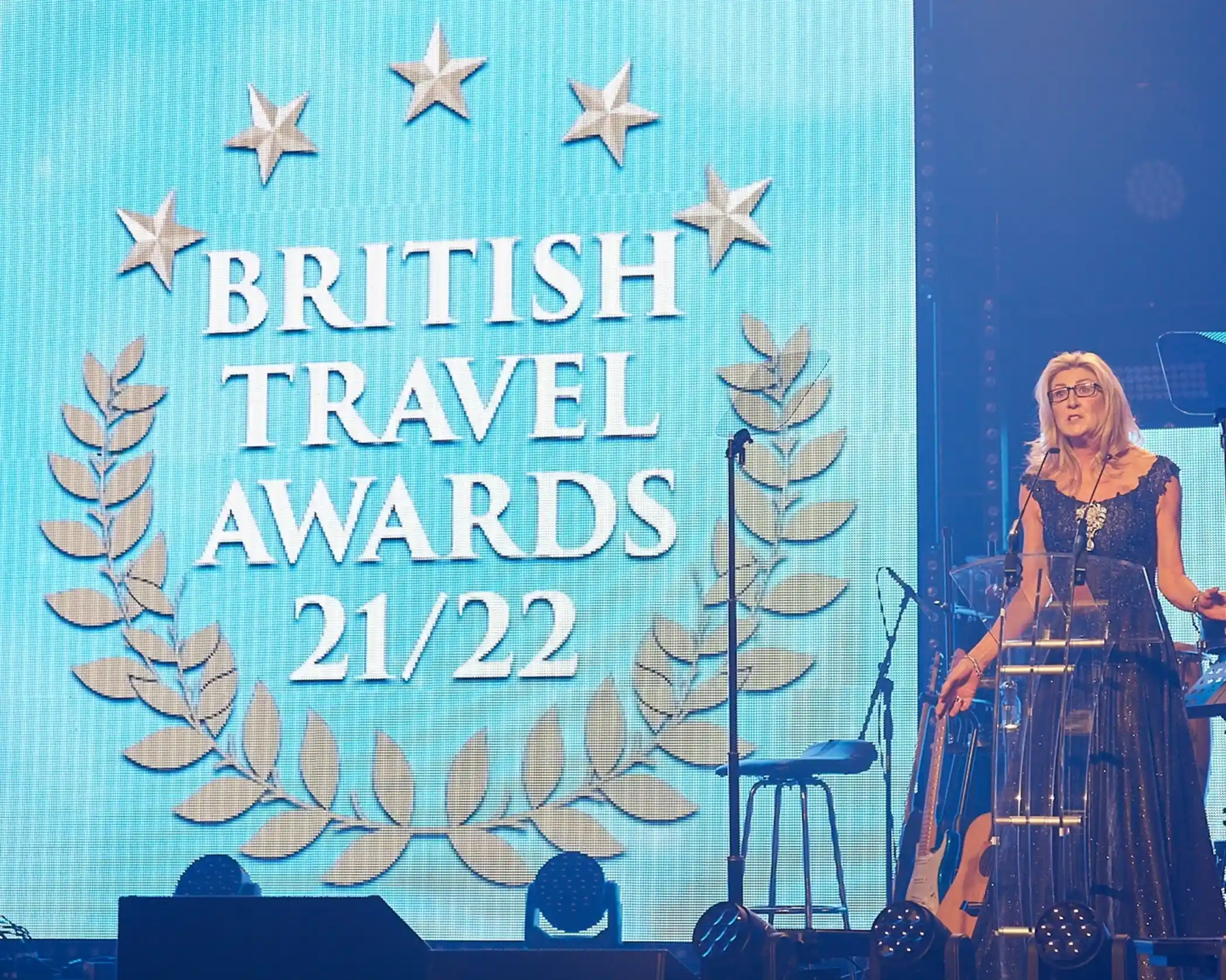 A woman in an elegant dark gown stands at a clear podium on stage, speaking at the British Travel Awards 2021/2022. Behind her is a large blue screen displaying the event logo with five stars, laurel wreaths, and the words 