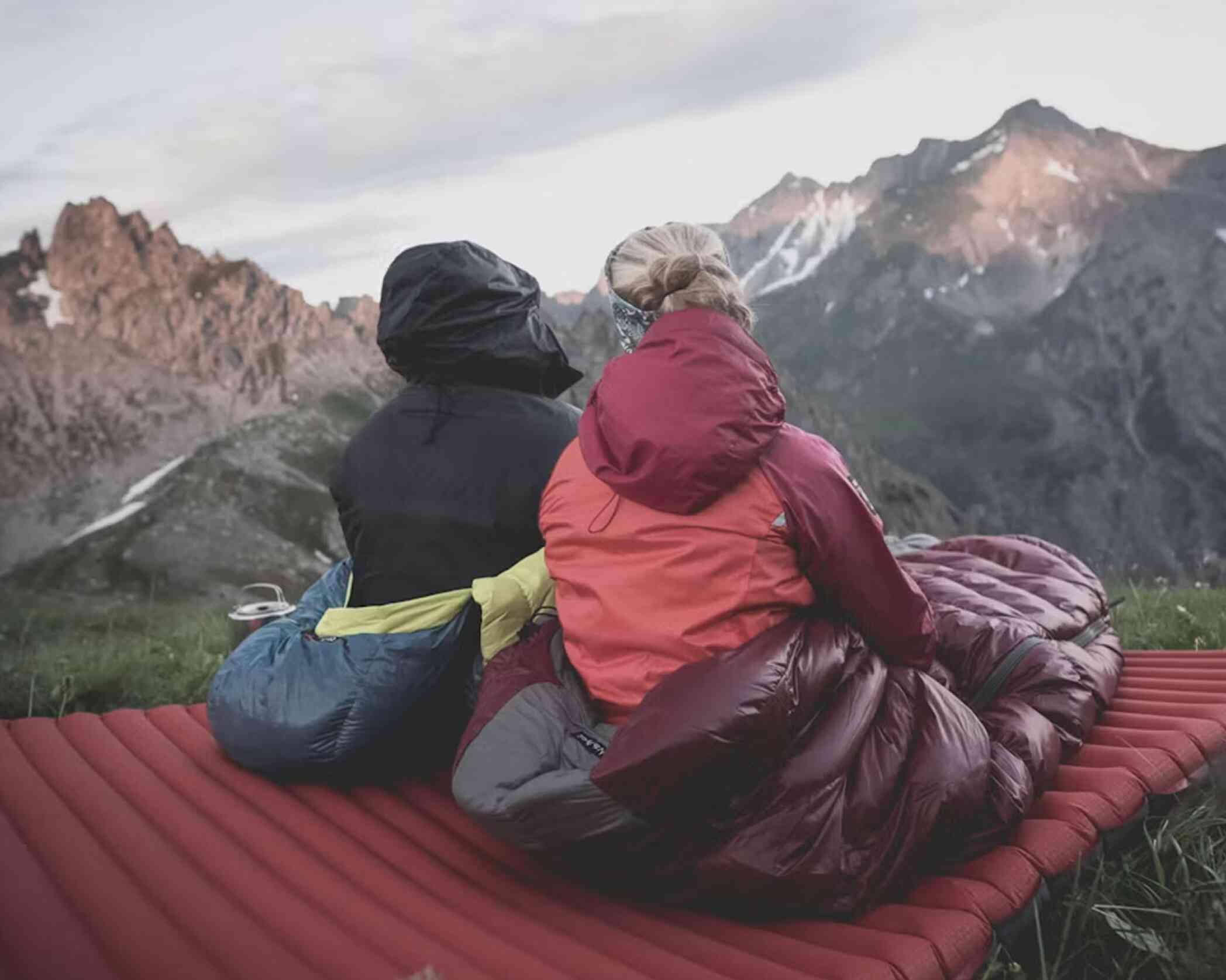 Two individuals sit side by side on a red camping mat, overlooking a mountainous landscape. They are wrapped in colorful sleeping bags, with peaks illuminated by the morning light. The sky is slightly cloudy, adding a serene atmosphere to the scene.