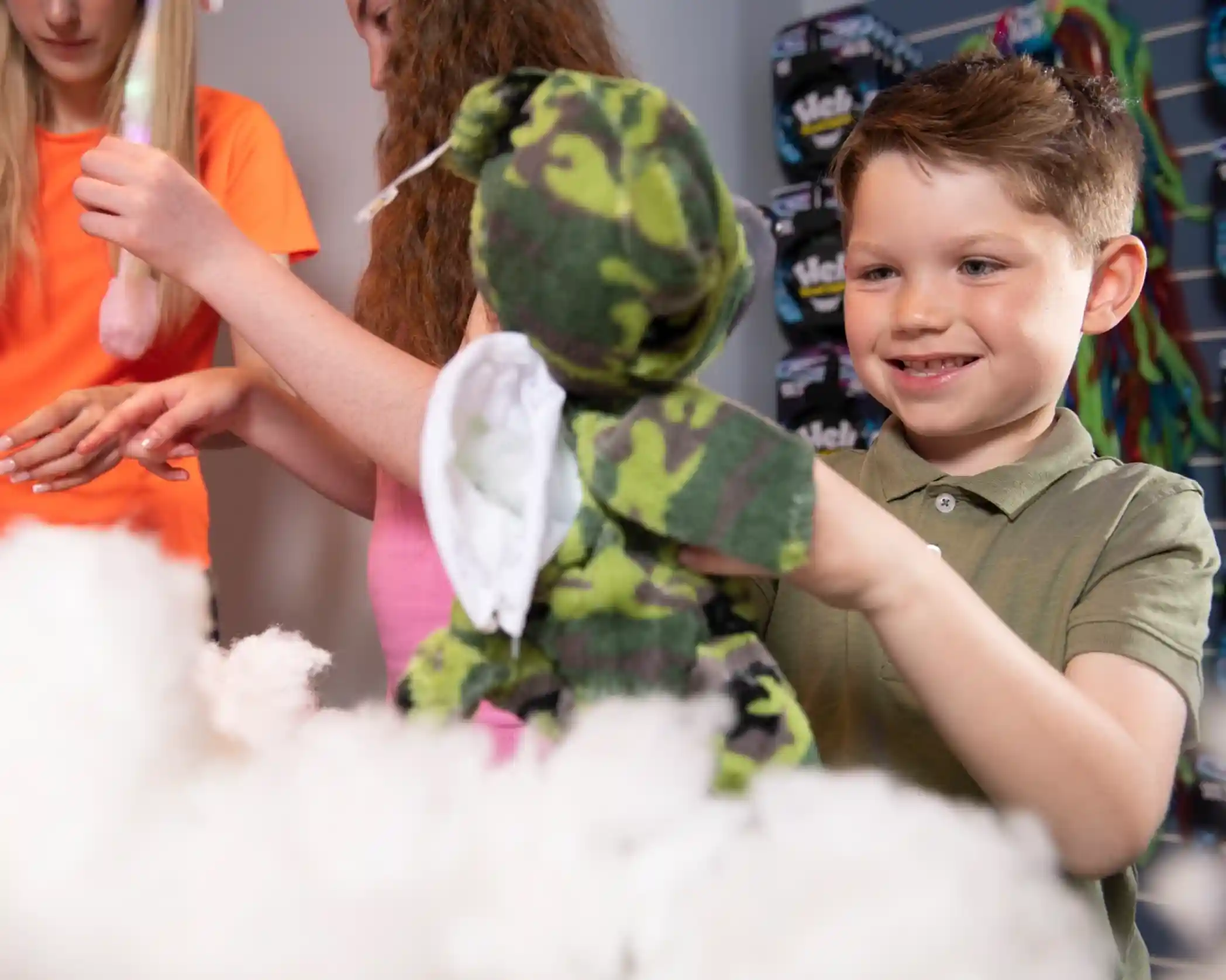 A young boy smiles while holding a camouflage teddy bear surrounded by fluffy white stuffing. Two girls are visible in the background, one holding a colorful object.