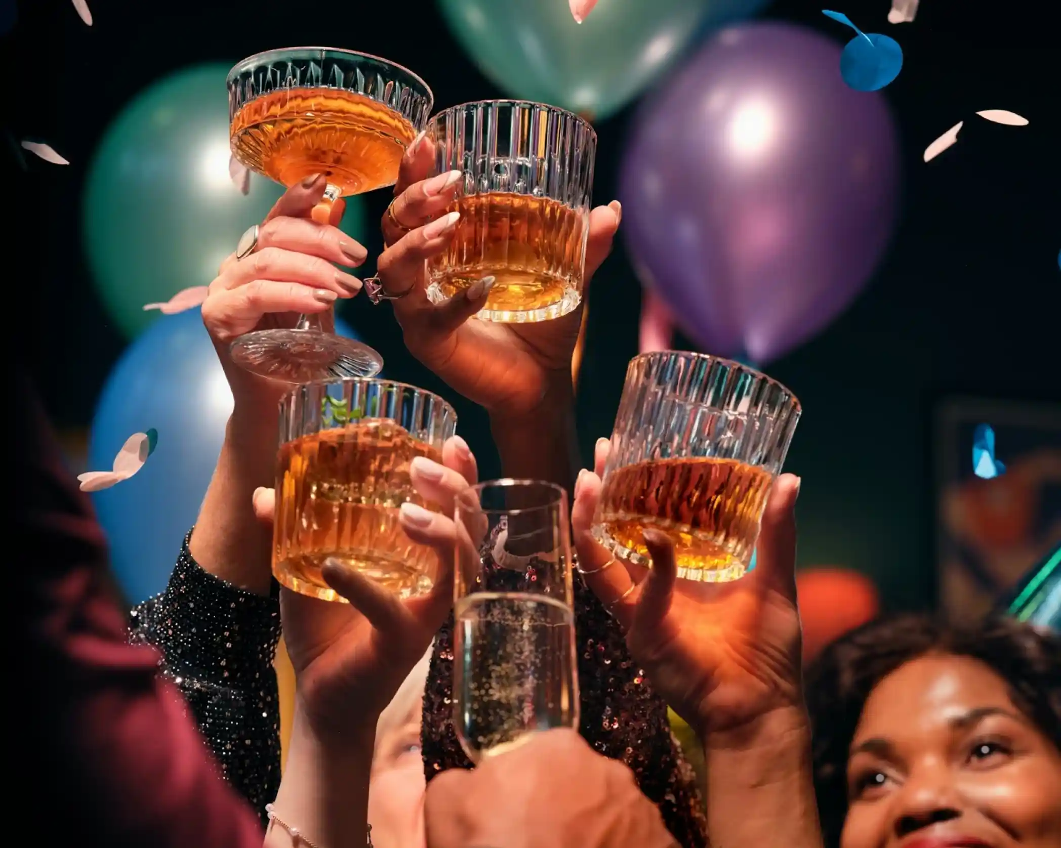 Hands hold up glasses filled with drinks in celebration, surrounded by colorful balloons and confetti.