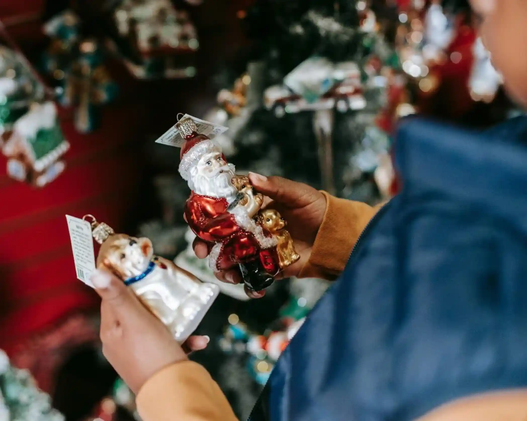 A person holds two Christmas ornaments, one depicting Santa Claus and the other a dog. They are surrounded by festive decorations in a shop.