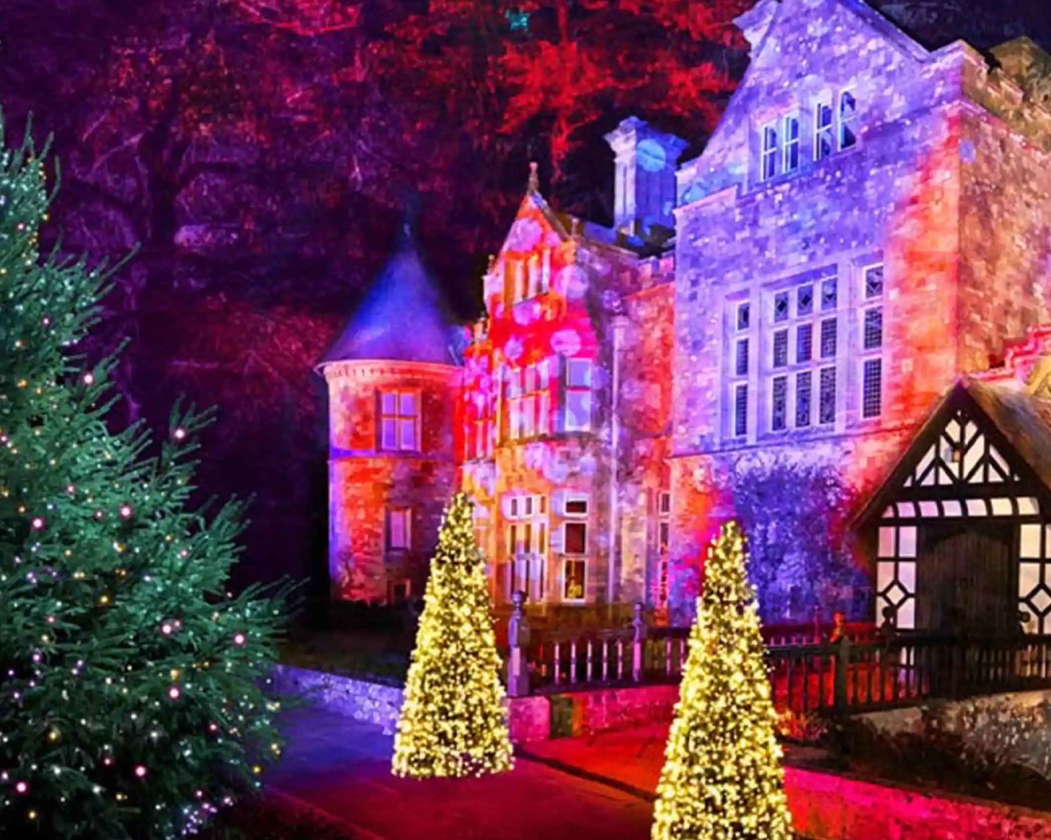 A beautifully illuminated stone mansion with colorful light projections decorating its facade. Two dazzlingly lit Christmas trees flank the entrance, with a festive atmosphere enhanced by vibrant colors in the surrounding landscape.