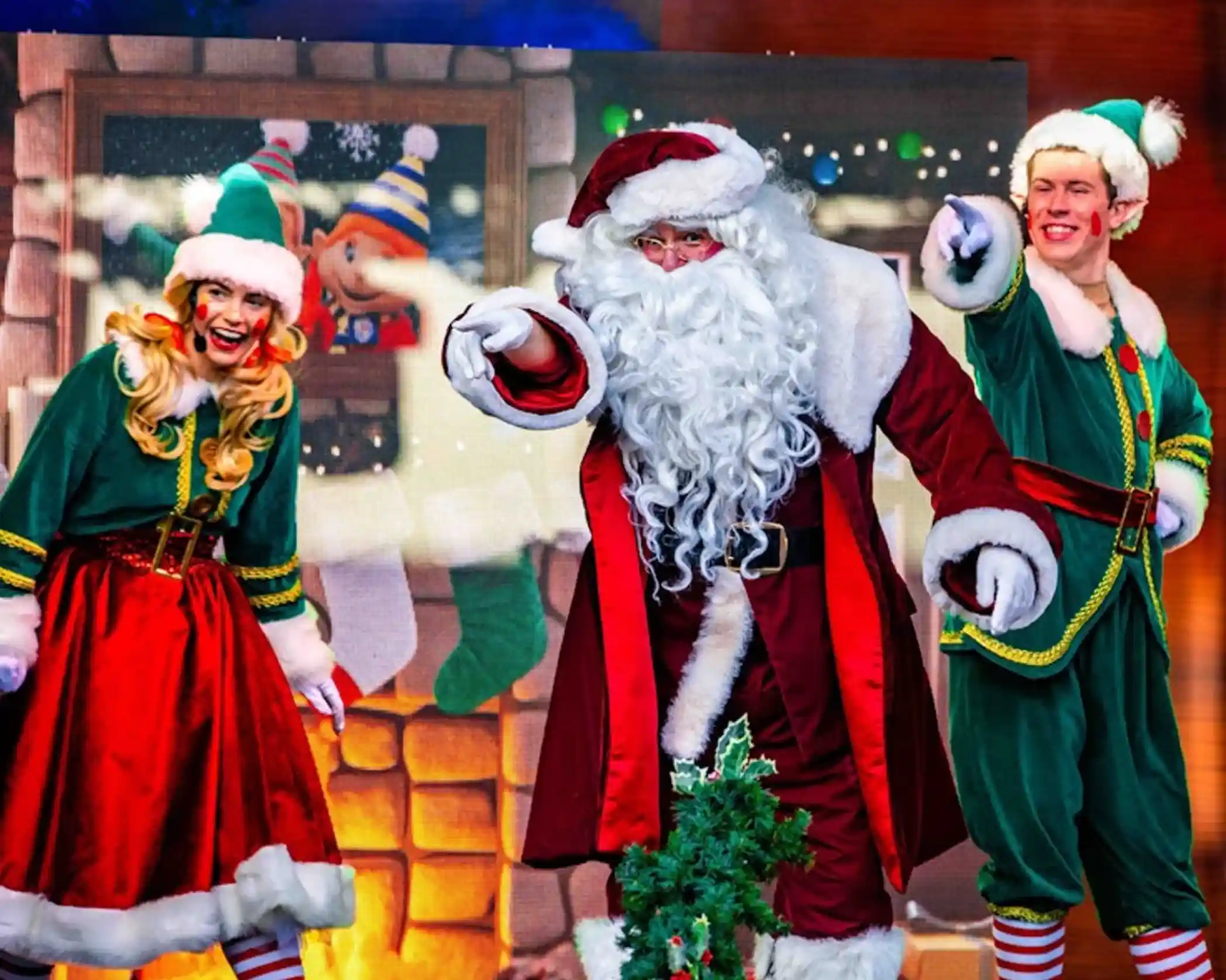 Santa Claus and two elves are performing on stage. Santa wears a traditional red suit and white beard, while the elves sport green outfits with red accents. They are all smiling and pointing, surrounded by a festive backdrop depicting a cozy holiday scene.