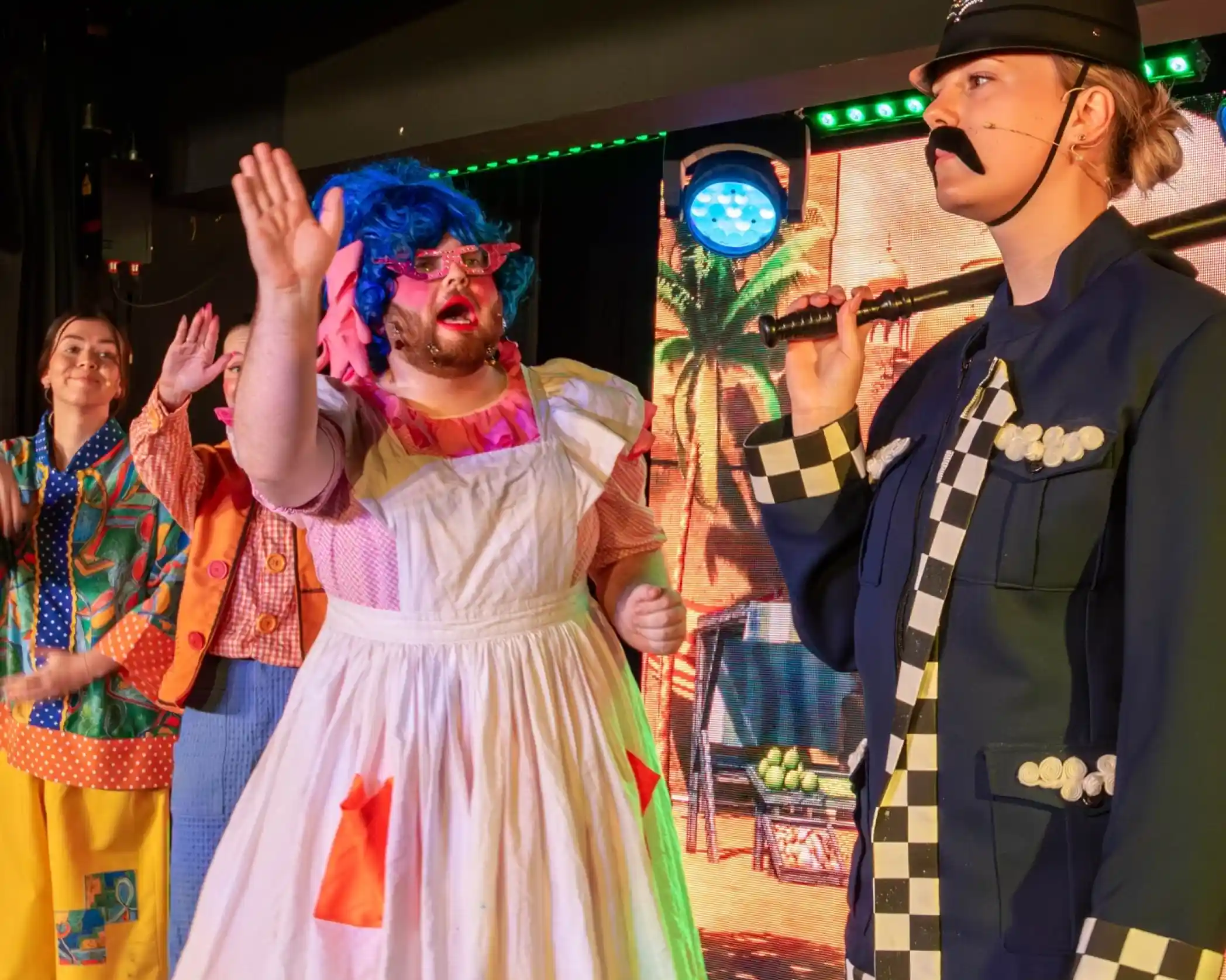 A performer in a colourful costume with blue hair and oversized glasses gestures animatedly while another performer dressed as a police officer observes. The background features colourful stage lighting and a tropical scene, with other cast members visible in vibrant outfits.