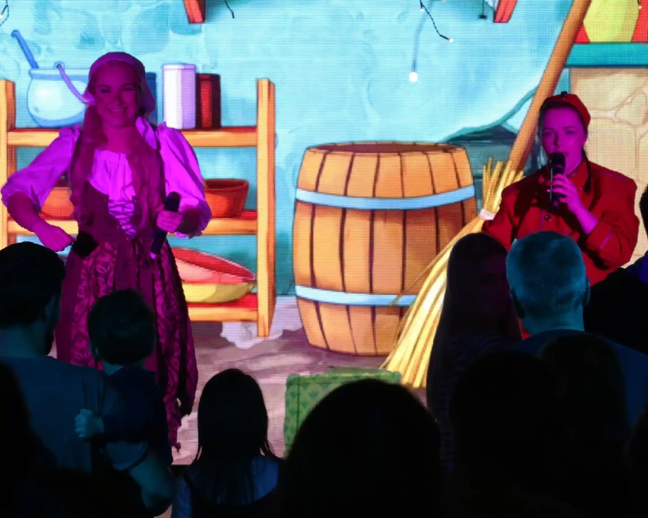 Two performers in colorful costumes are singing on stage, with a vibrant animated backdrop featuring a cabin interior. The audience is watching, silhouetted in the foreground.