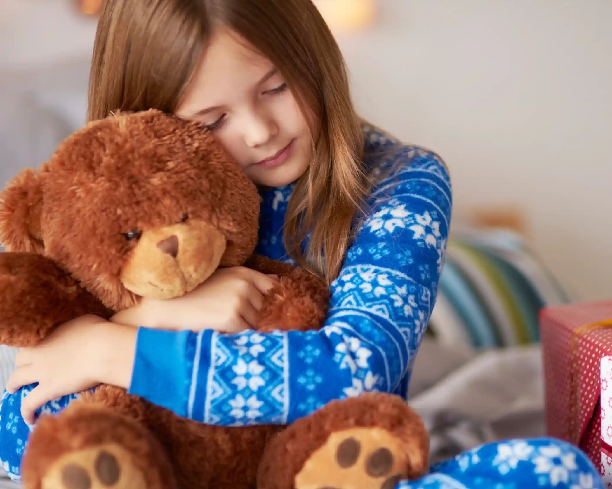 A girl in blue patterned pajamas hugs a large brown teddy bear, smiling gently with her eyes closed. Wrapped presents are visible in the background.