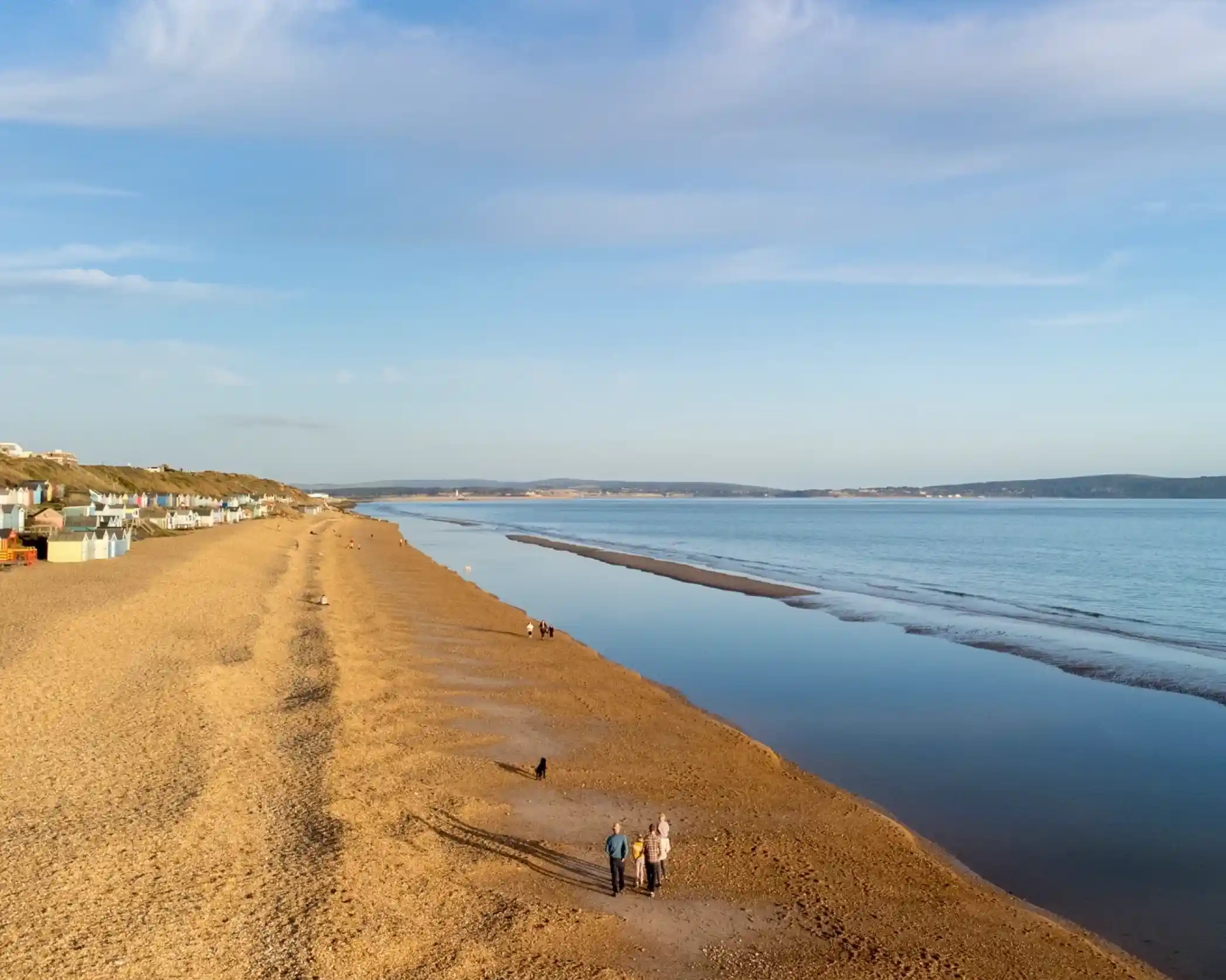 A serene beach scene with golden sand stretching along the shore. A couple walks hand-in-hand, their shadows cast on the sand. Calm waves lap at the beach, with colorful beach huts visible in the background. The sky is clear with soft, wispy clouds.