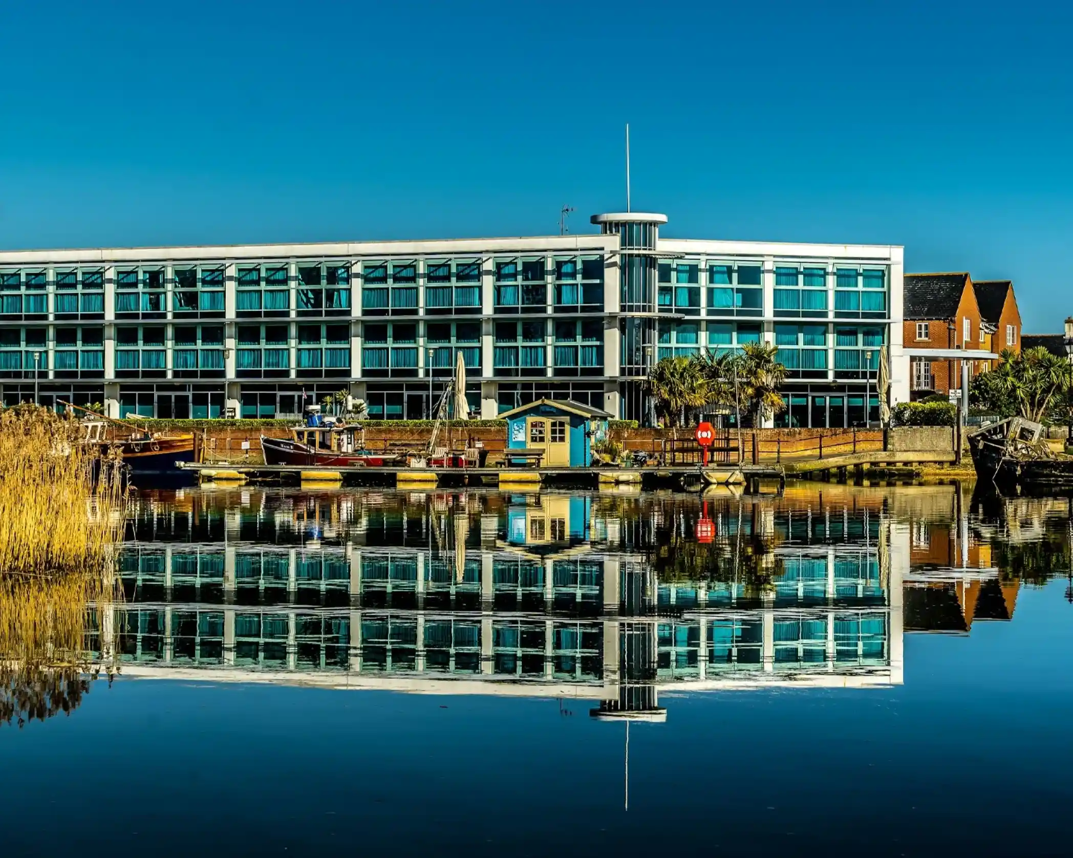 Modern waterfront building with large glass windows reflecting in calm water, alongside boats and a small shed. Clear blue sky above with some vegetation in the foreground.