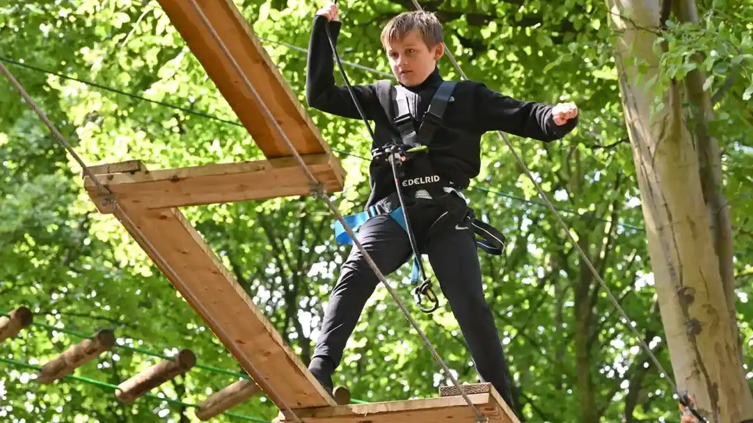 A child navigates a high ropes course, balancing on wooden beams while secured in a harness. Lush green trees surround the scene, creating a vibrant outdoor setting.