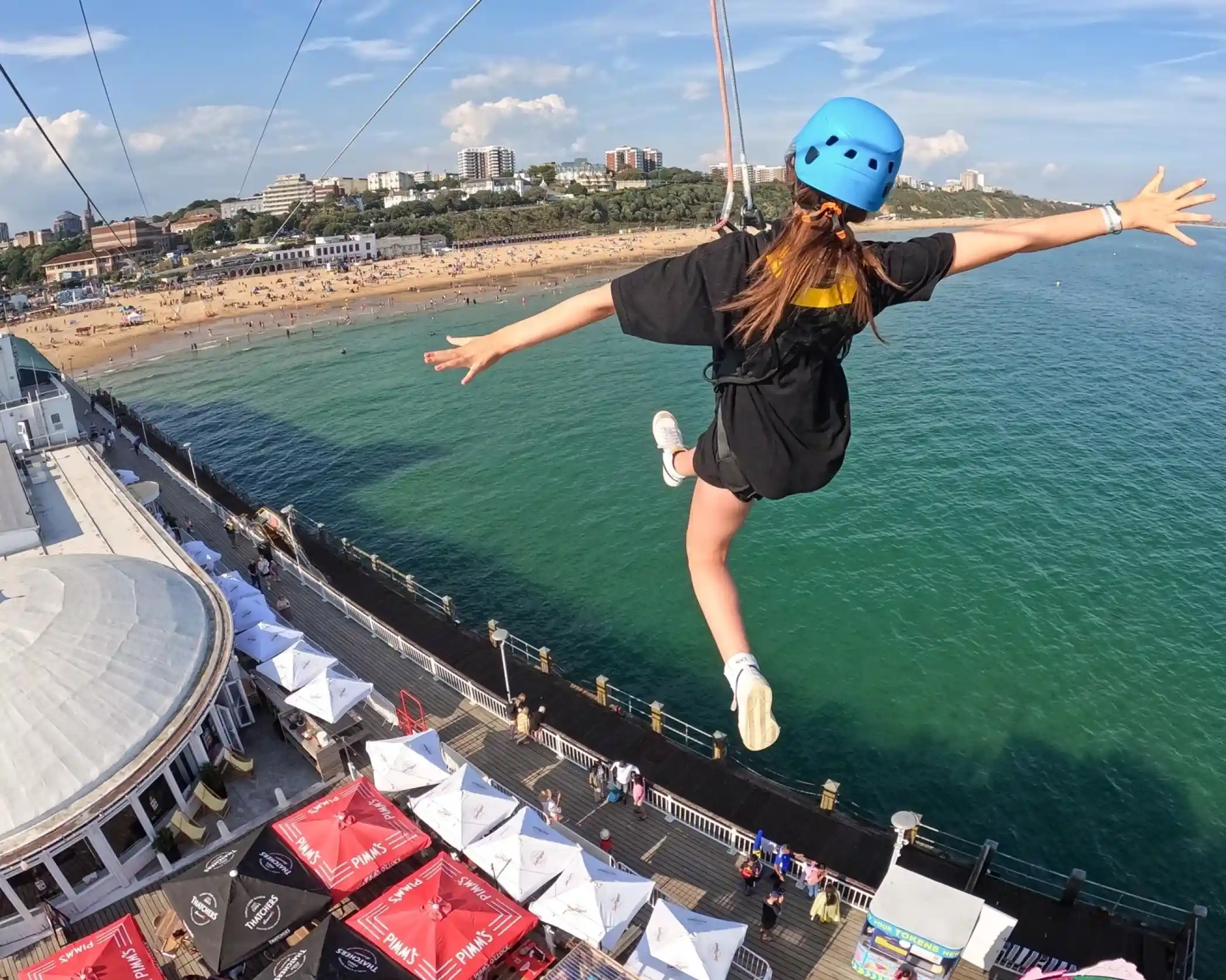A person in a blue helmet and black shirt is zip-lining above a beach with a crowd of people, colorful umbrellas, and ocean views. The scene captures a sunny day with clear skies.