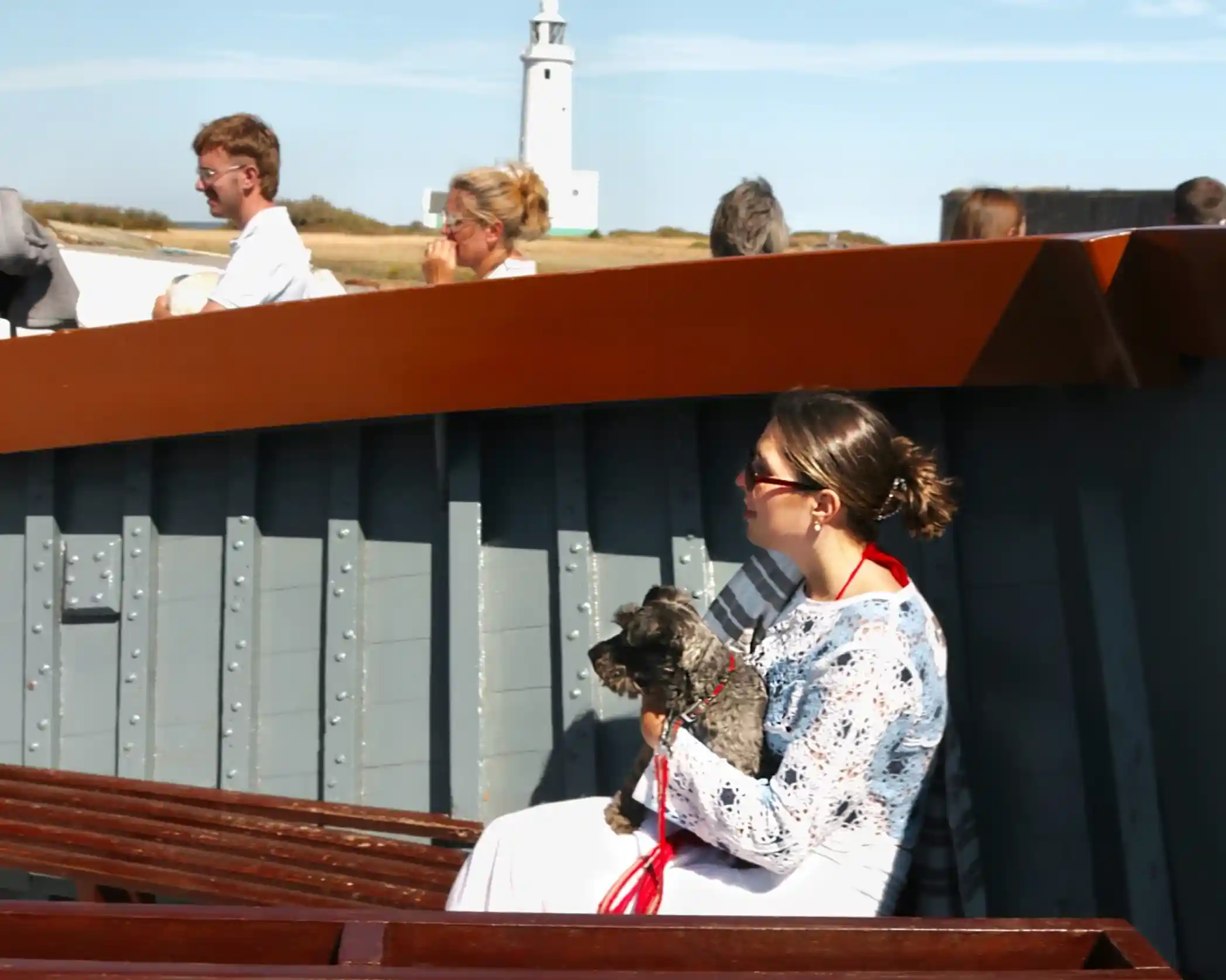 A woman holds a small dog while sitting on a boat, wearing a white dress and sunglasses. In the background, a lighthouse and people are visible enjoying the sunny day.