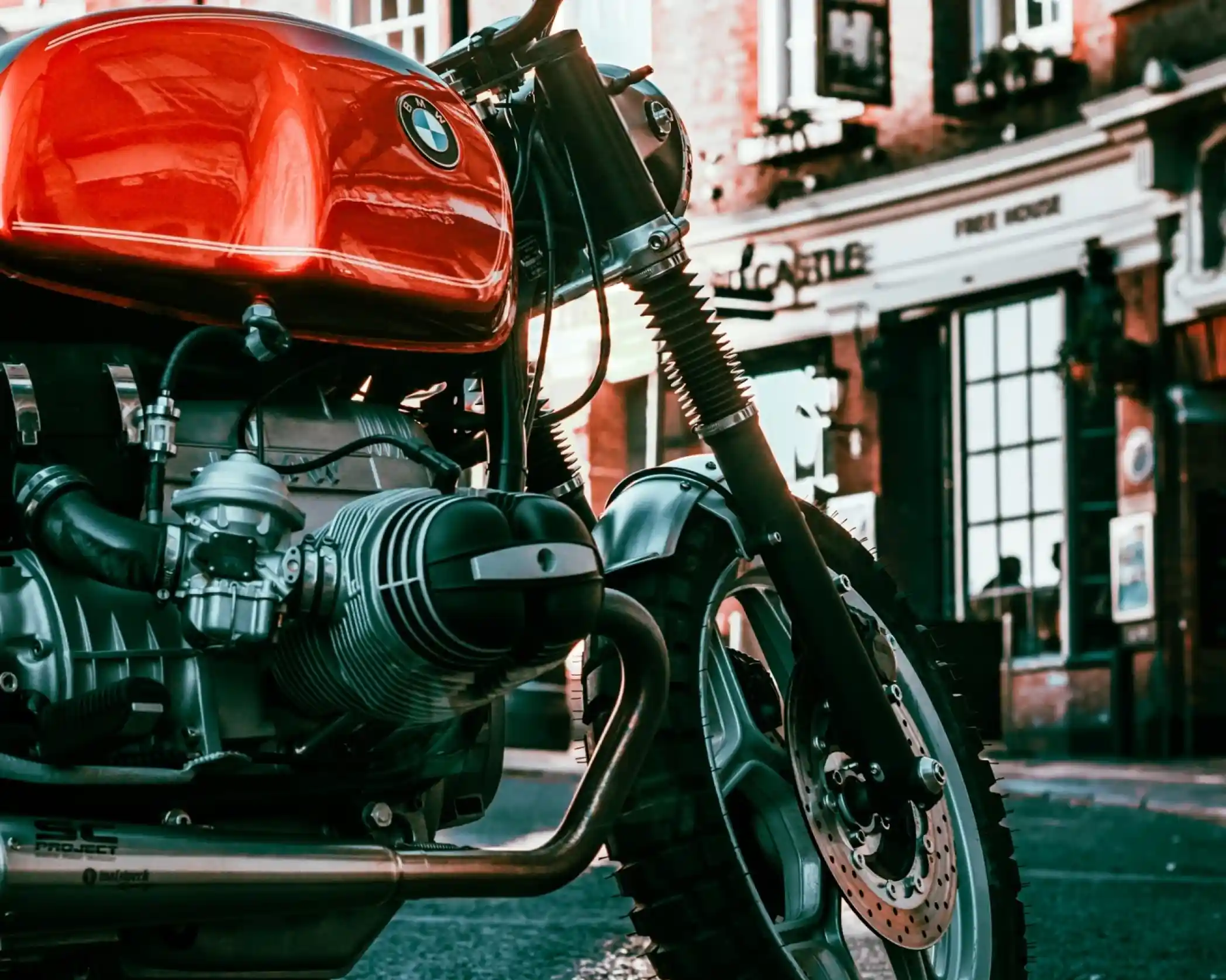 A close-up view of a red BMW motorcycle parked on a street. The bike features a vintage design with a prominent engine and chrome details. In the background, a pub building is visible, adding to the urban atmosphere.