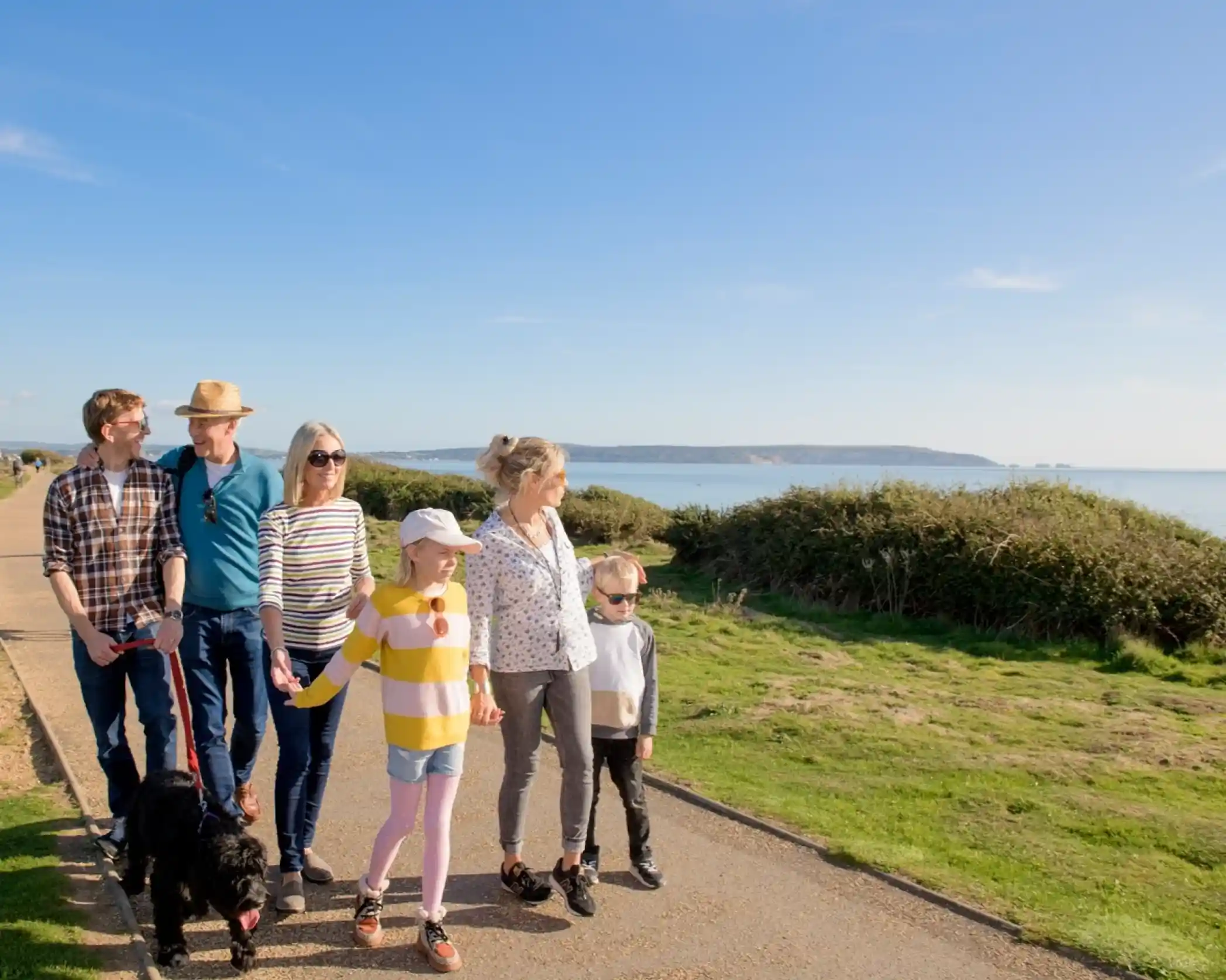 A group of six people walks along a coastal path, enjoying a sunny day. They include adults and children, with one person holding a black dog. The background features greenery and a view of the water.