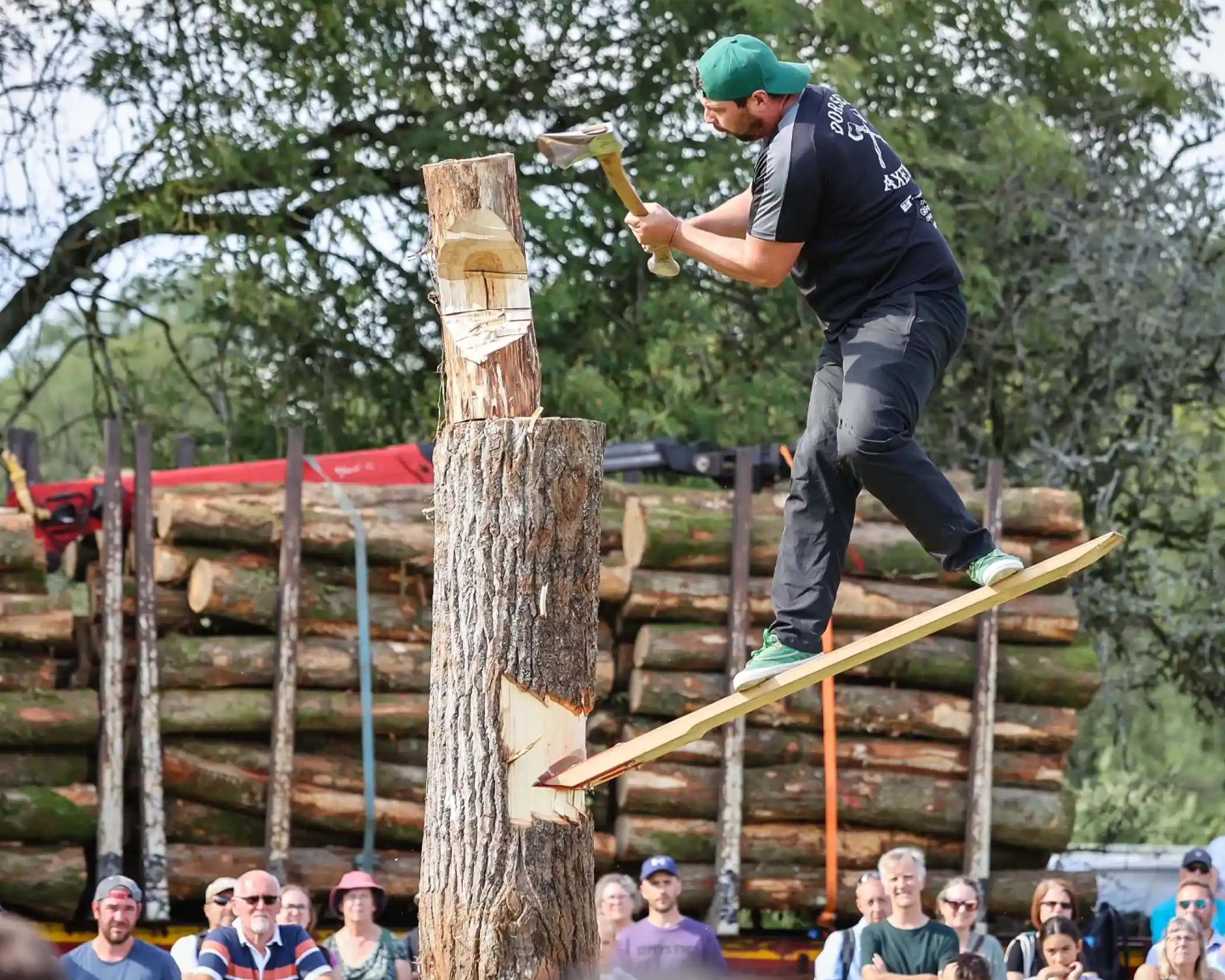 A lumberjack balances on a narrow board while swinging an axe at a wooden log. In the background, a stack of logs and an audience watches the competition.