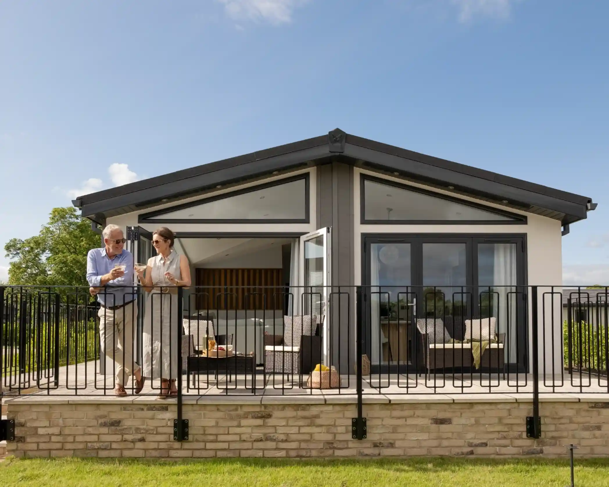 A couple stands on a deck in front of a modern house with large windows. The man gestures as he talks, while the woman leans against a railing, smiling and listening. The scene is sunny, surrounded by greenery.