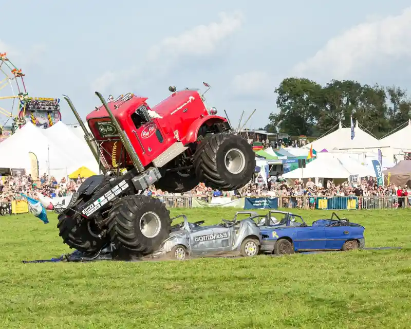 A red monster truck with oversized tires jumps over two crushed cars during a performance in a field filled with spectators and event tents in the background.
