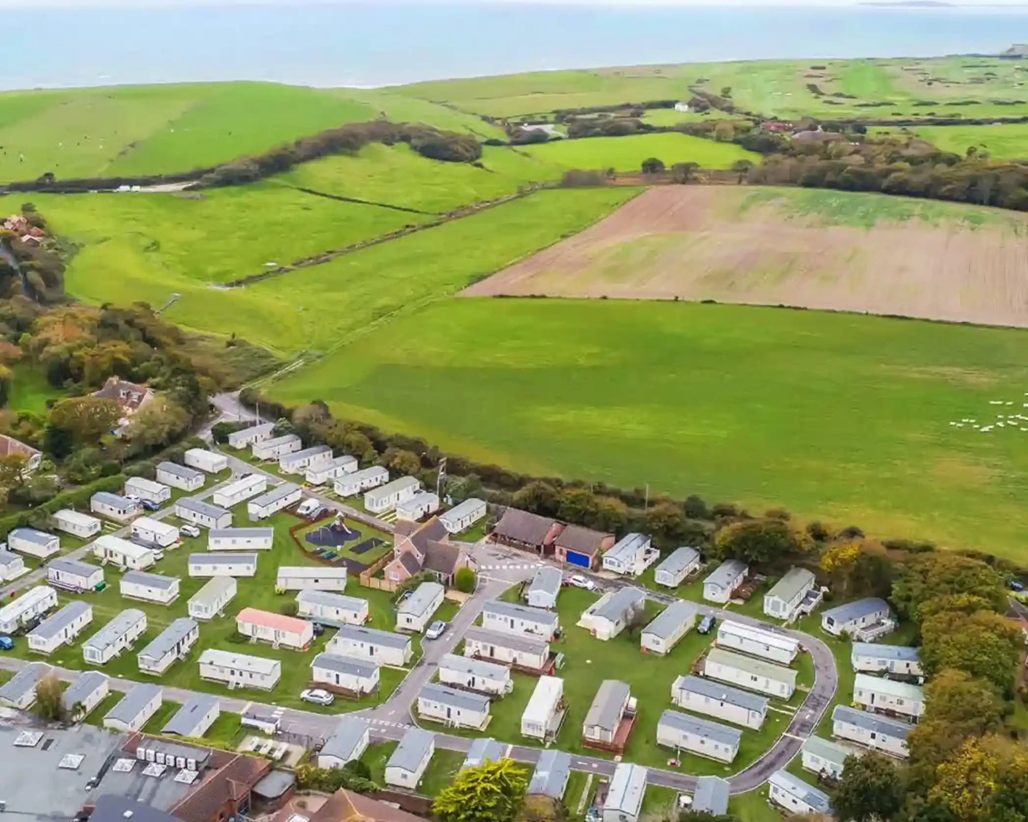 Aerial view of a caravan park with neatly arranged caravans, surrounded by lush green fields and hills, overlooking the sea in the background.