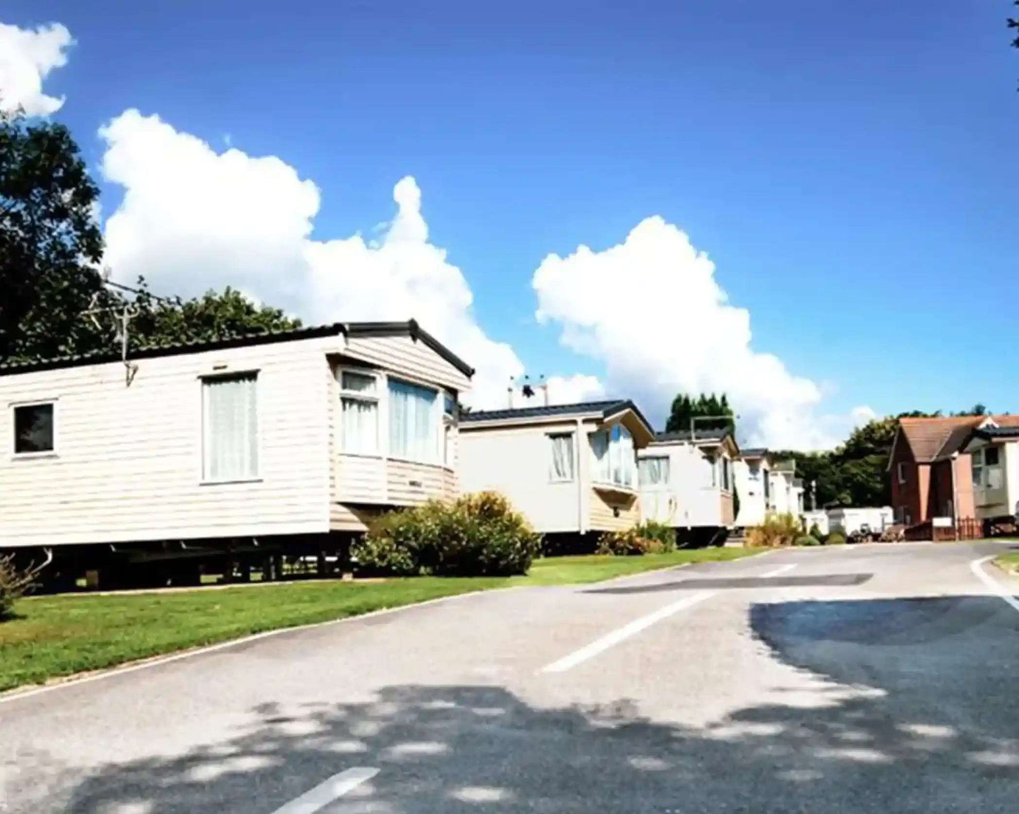 A row of holiday caravans along a quiet road, surrounded by grass and trees, with a bright blue sky and fluffy white clouds in the background.