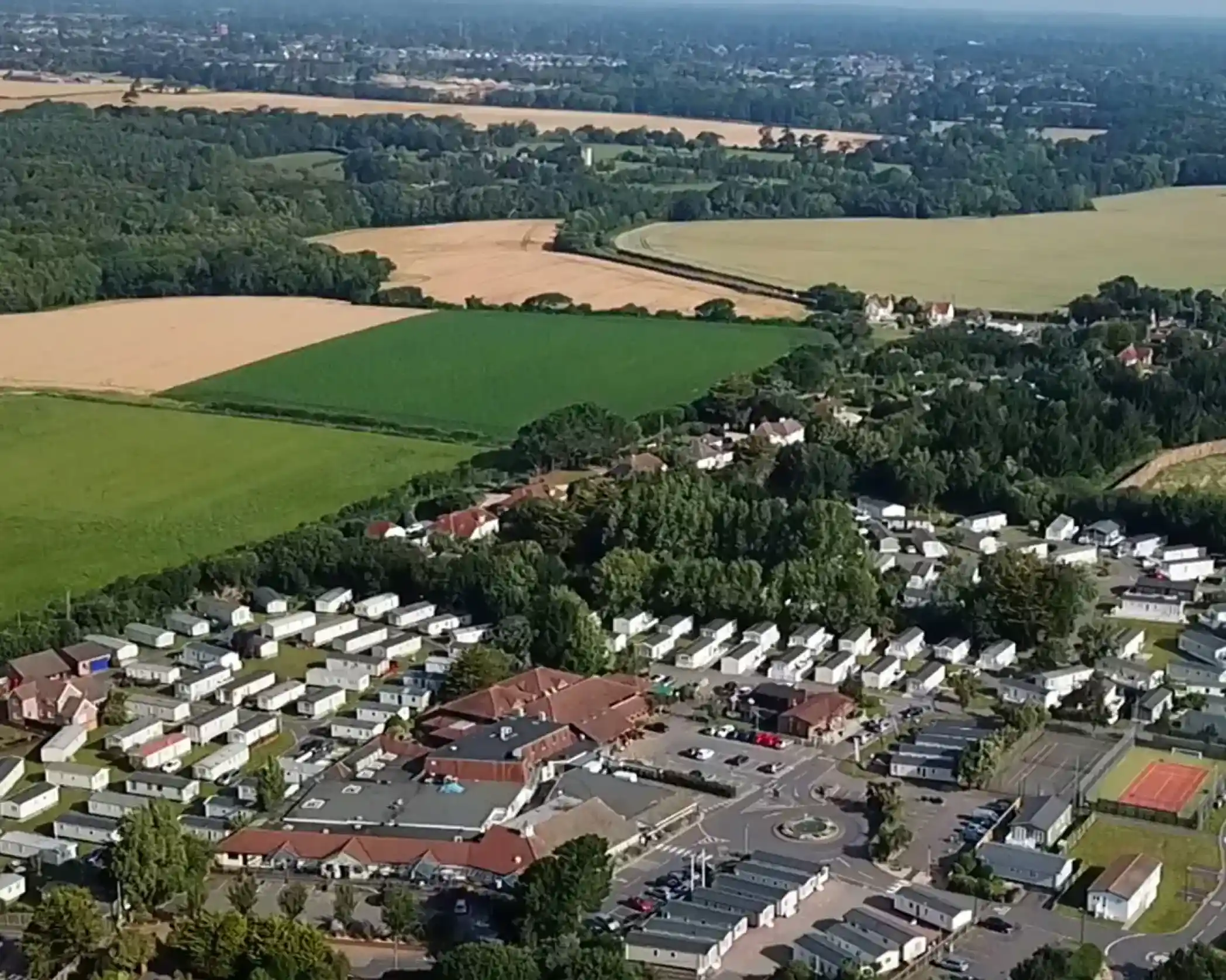 Aerial view of a rural landscape featuring a cluster of mobile homes, lush greenery, fields, and distant hills under a clear blue sky. The area shows a mix of residential buildings, sports facilities, and open farmland.