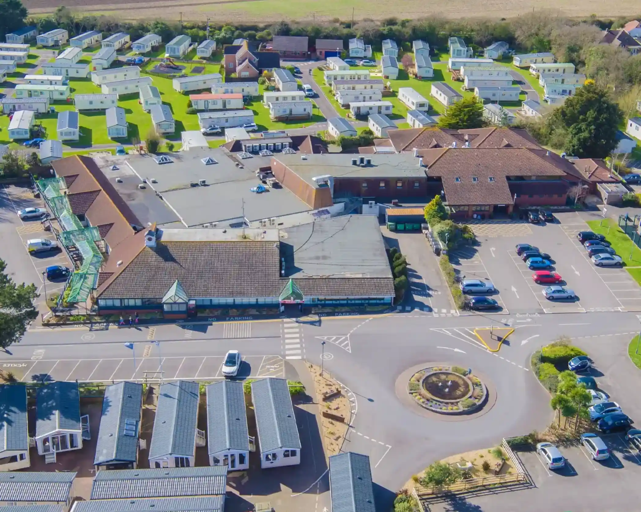 Aerial view of a caravan park featuring numerous mobile homes arranged neatly on green lawns. In the center, there is a commercial building with a parking area, surrounded by vehicles. A roundabout is visible in the foreground, leading into the park.