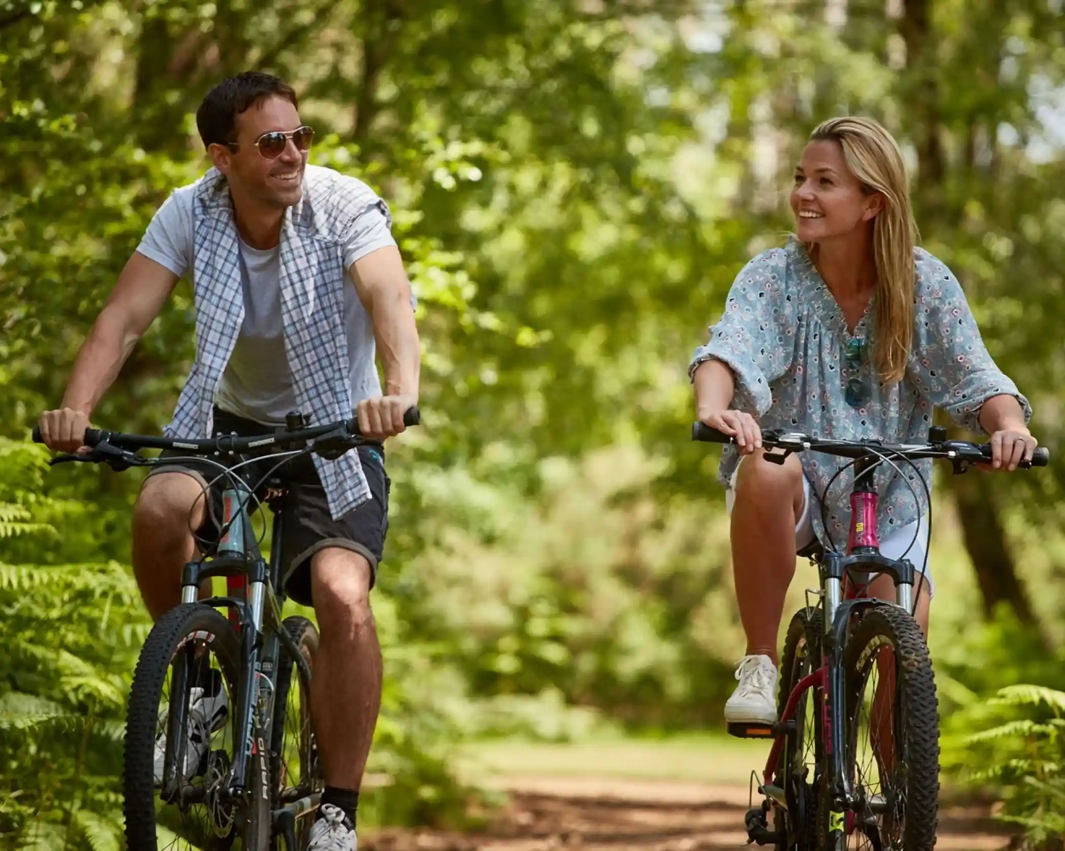 A man and a woman ride bicycles on a forest path, smiling at each other amidst lush greenery. The man wears sunglasses and a casual shirt, while the woman wears a light, patterned dress and sneakers.