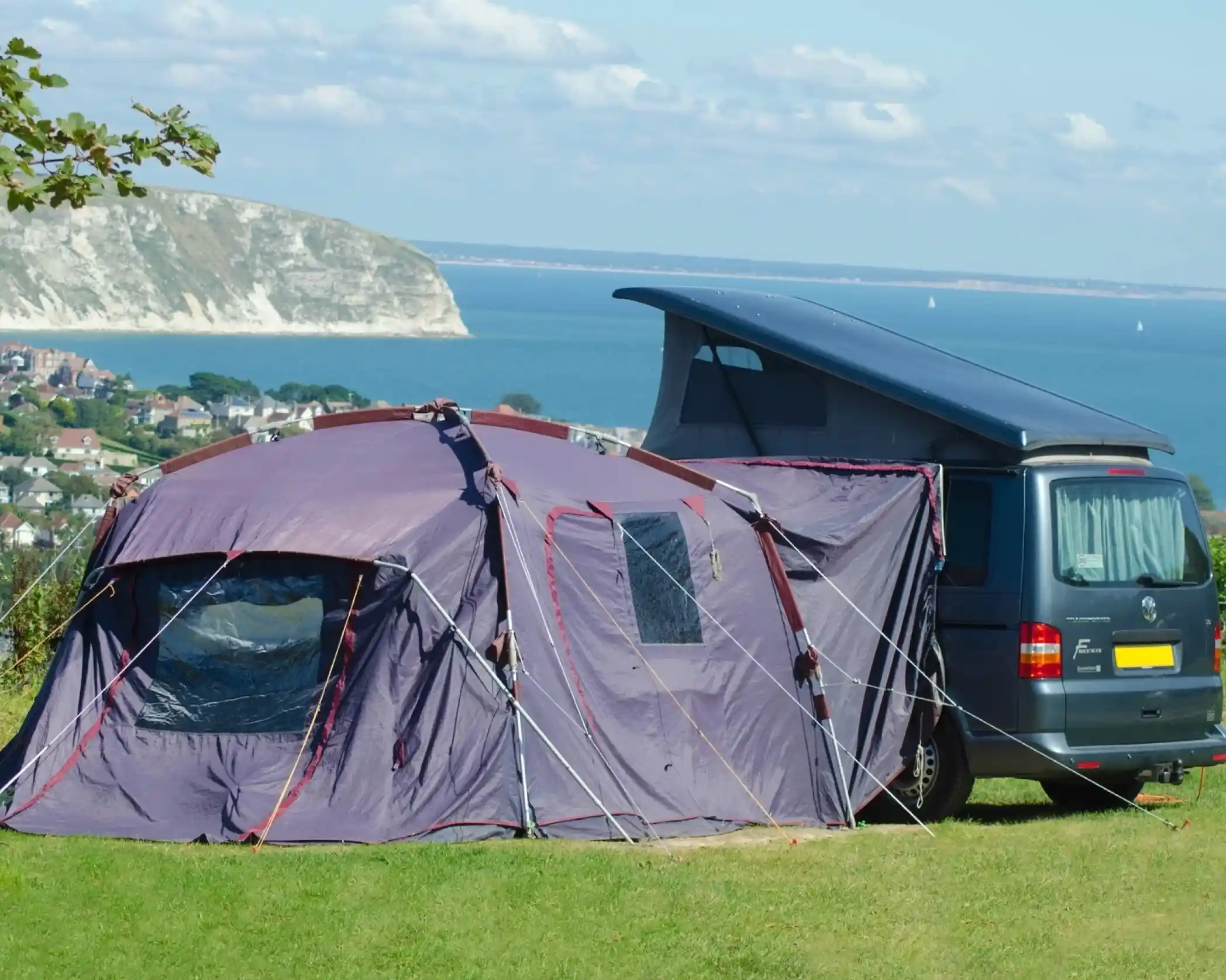 A camper van parked next to a large tent on a grassy hill overlooking a scenic coastline with distant cliffs and a blue ocean.