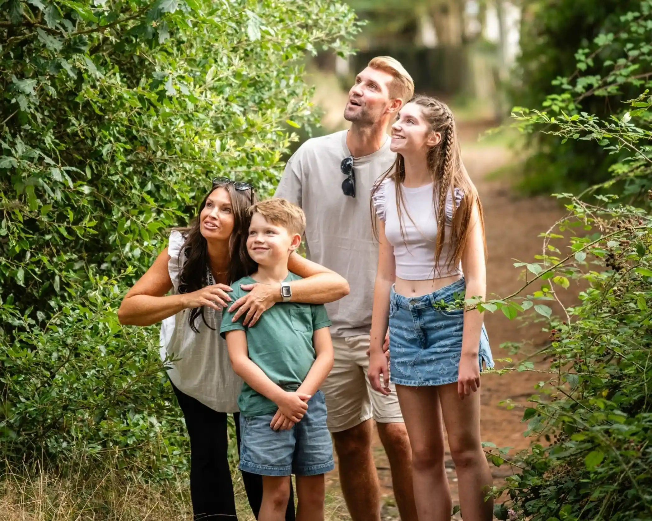 A family of four stands on a woodland path, looking up in awe. The mother, wearing a light top, points towards something interesting. A young boy in a green shirt stands beside her, while a teenage girl in a white top and denim skirt gazes up as well. The father, casually dressed, watches with a surprised expression. Lush greenery surrounds them.