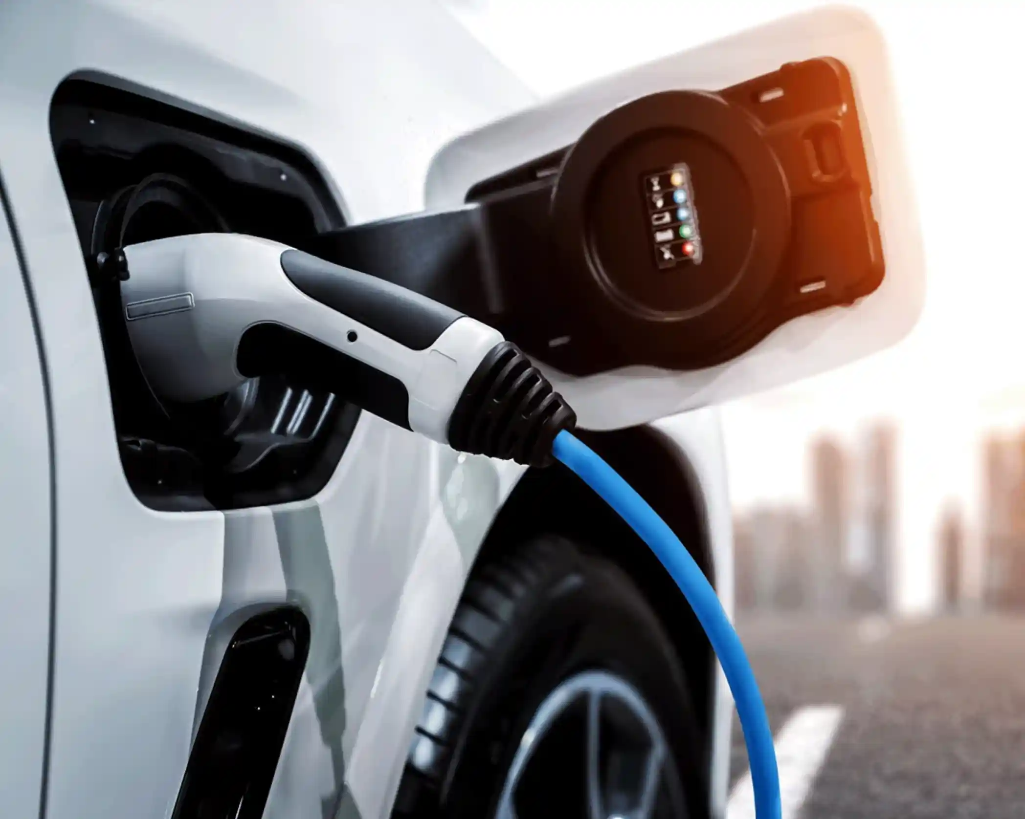 A close-up of an electric vehicle charging with a blue charging cable connected to the charging port. The background shows an urban landscape with blurred buildings.