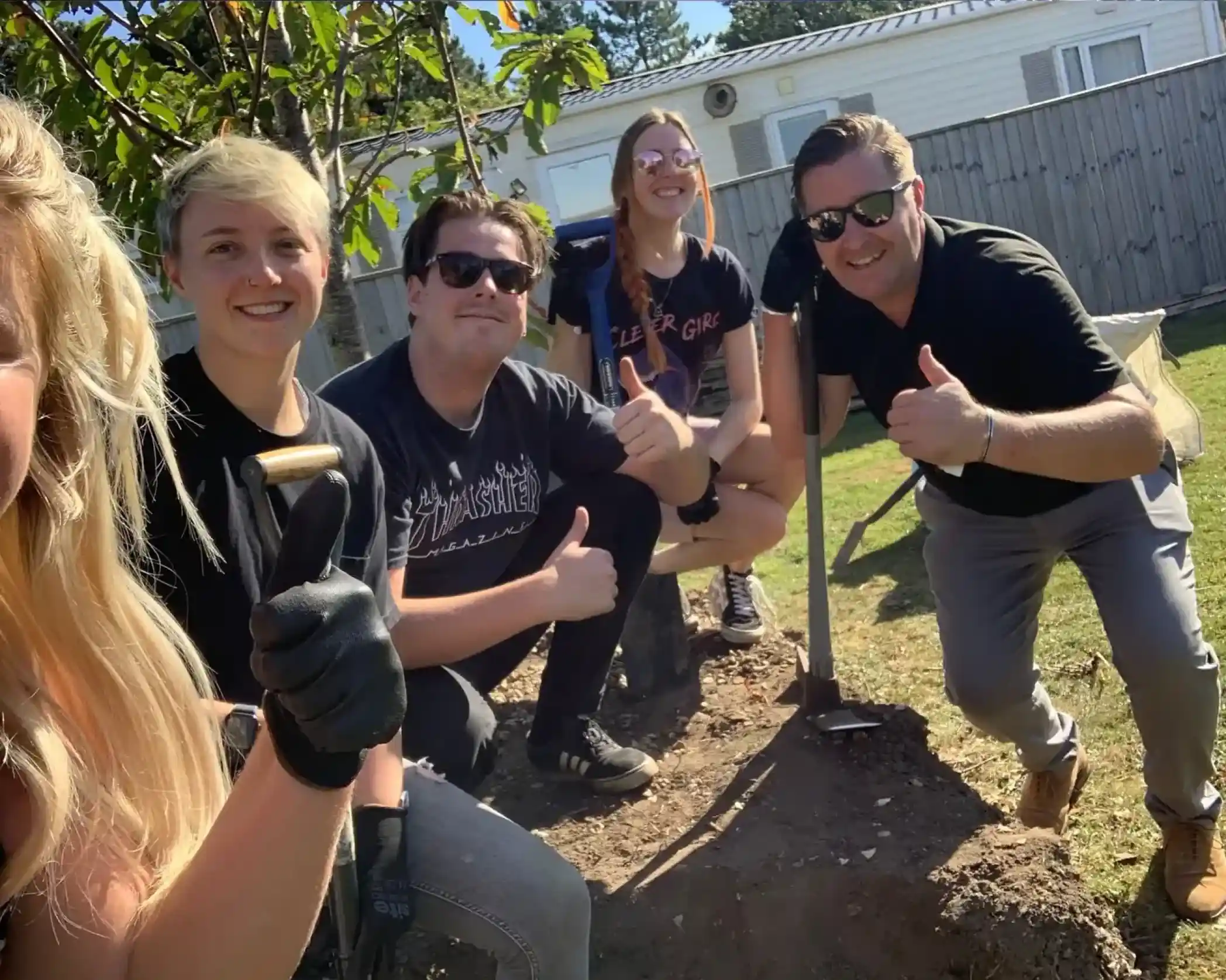 A group of five people outdoors, smiling and giving thumbs up while standing around a freshly dug hole. They are dressed casually, with gloves on some hands, and a spade is leaned against a tree. Sunlight brightens the scene, indicating a warm day.
