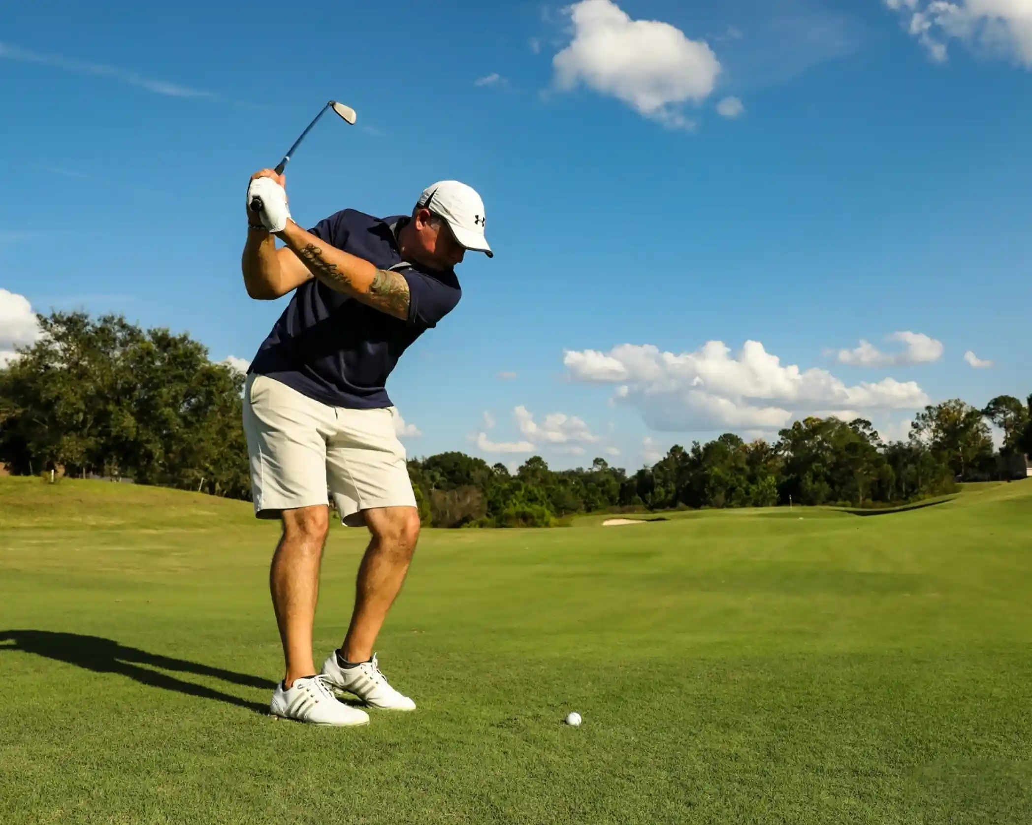 A man in a navy shirt and white cap swings a golf club on a sunny day. He stands on a lush green fairway, preparing to hit a golf ball. Fluffy white clouds float in a bright blue sky.