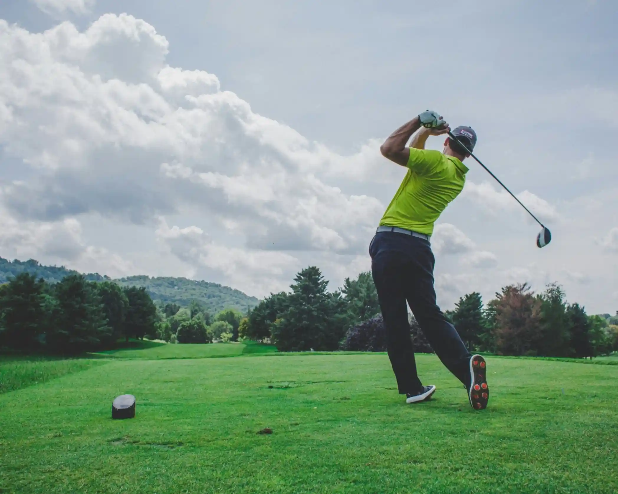 A golfer in a bright yellow shirt swings a driver on a lush green course, with rolling hills and clouds in the background.