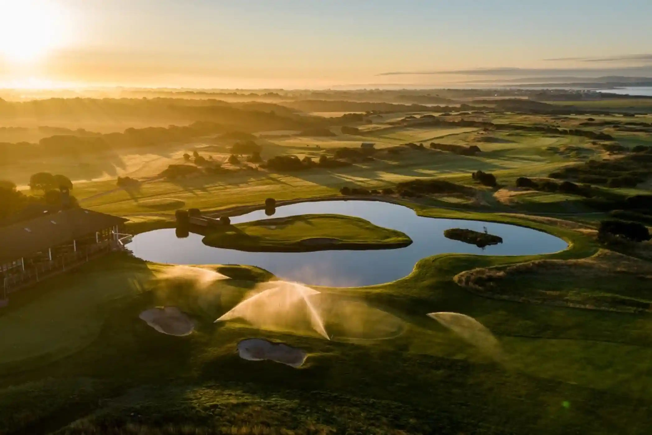 A picturesque golf course at sunrise, featuring lush green fairways, a tranquil pond, and mist rising from the ground. Soft sunlight illuminates the landscape, creating a serene atmosphere.