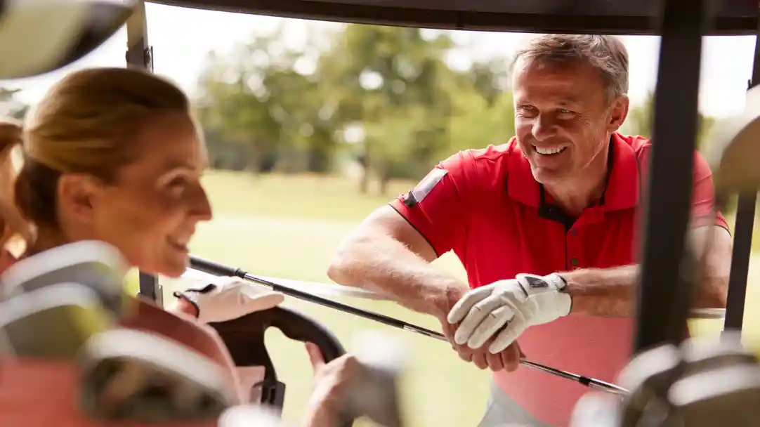 A man in a red polo shirt smiles while leaning on a golf cart, engaging with a woman who is seated in the cart, wearing golf gloves. Green grass and trees are visible in the background.